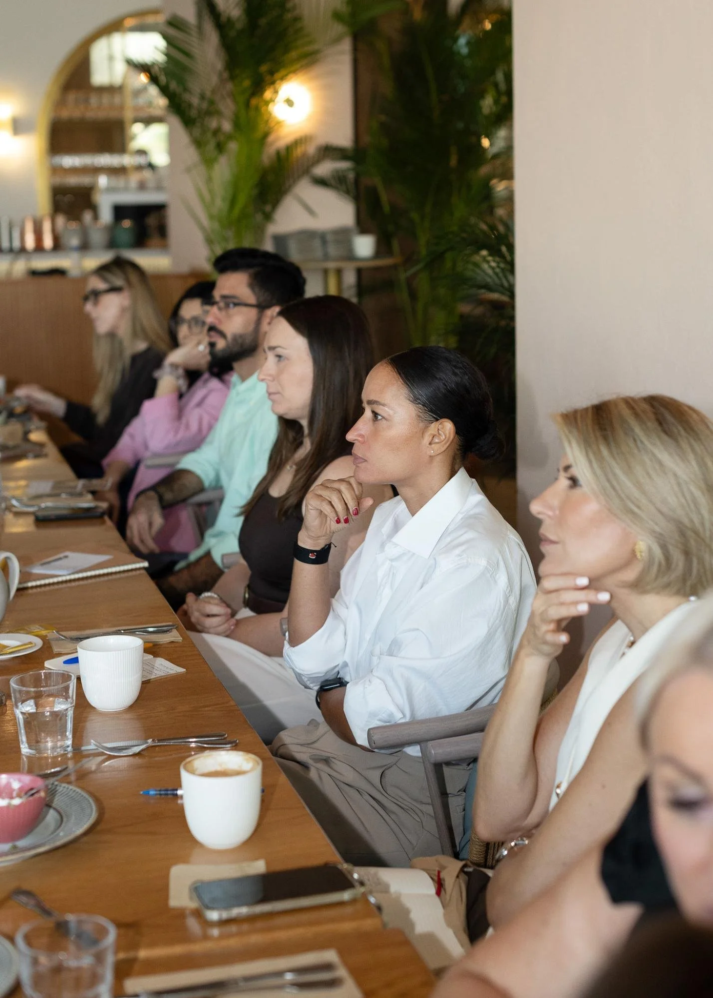 People sitting at a long wooden dining table in a restaurant, paying attention to a presentation or speaker. The table has cups, plates, utensils, and a notebook. The background has plants and warm lighting.