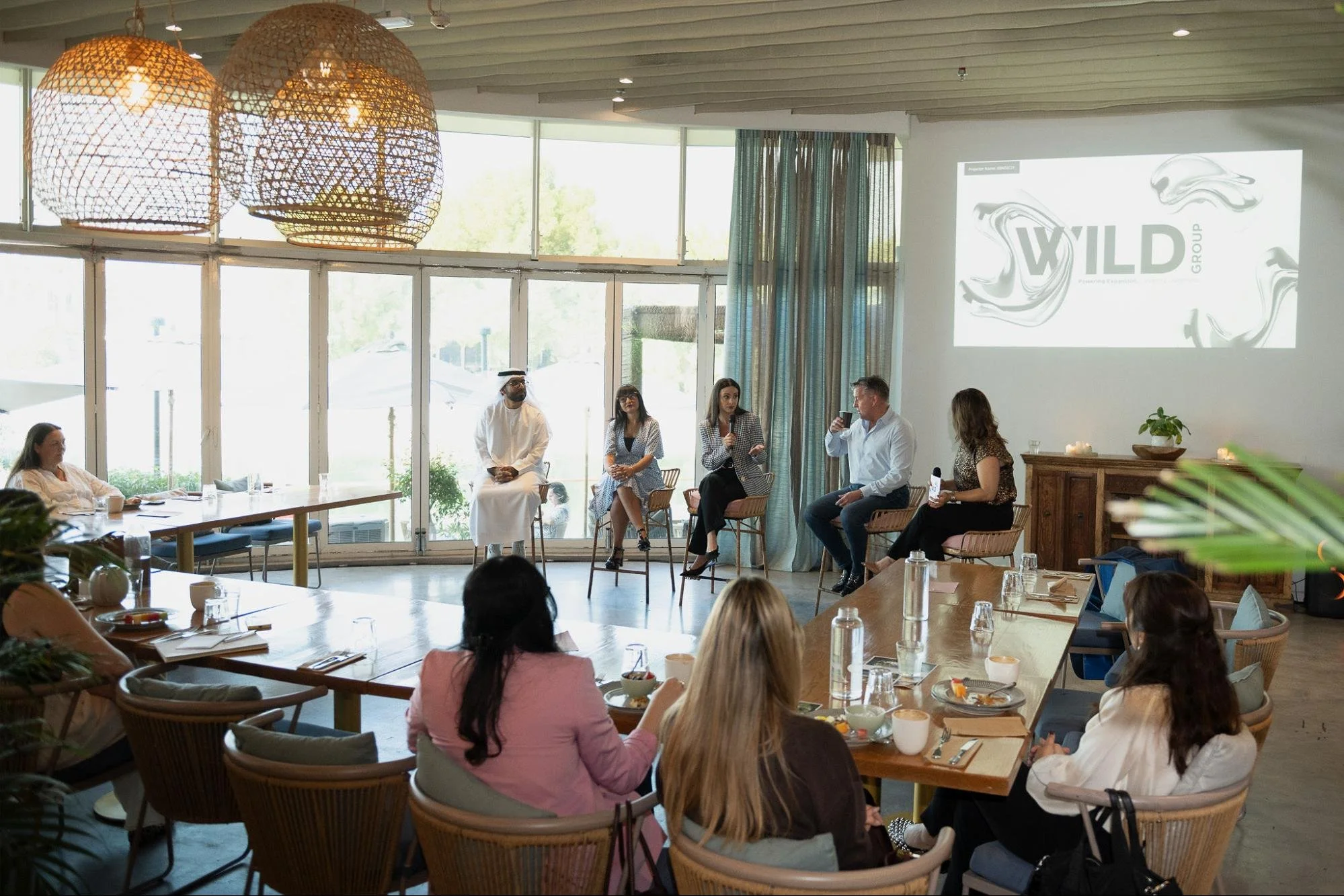 People sitting on stage during a panel discussion in a bright, modern room with large windows, hanging wicker lights, and a projector screen displaying a logo that reads 'WILD GROUP'.
