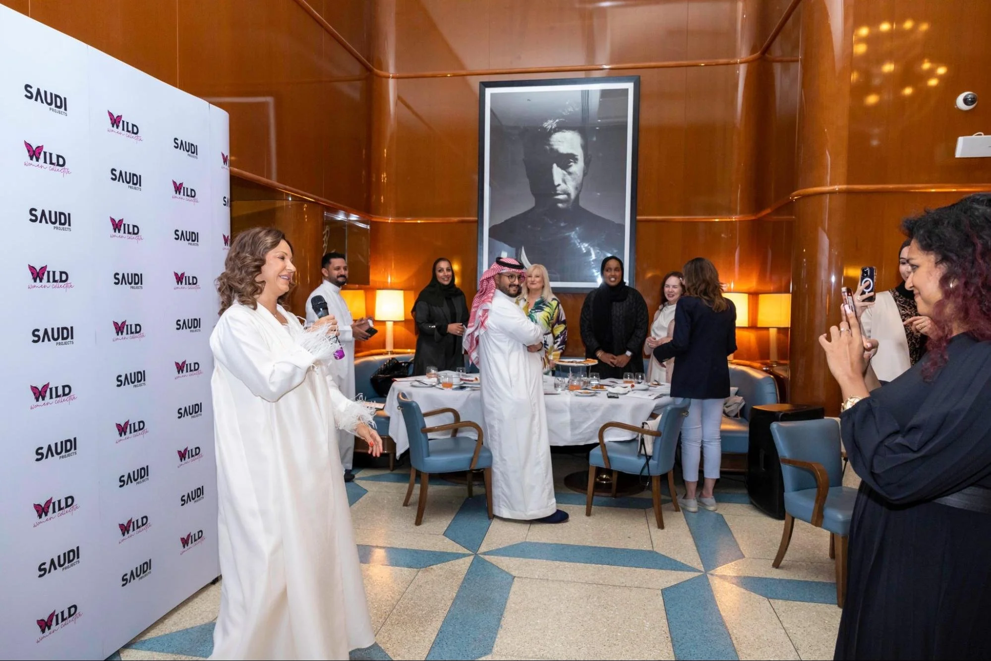 Group of diverse women and men attending a women’s empowerment event in a conference room with wood panel walls and a large black and white portrait of a woman on the wall. Some are taking photos, talking, or smiling. A woman in a white traditional d