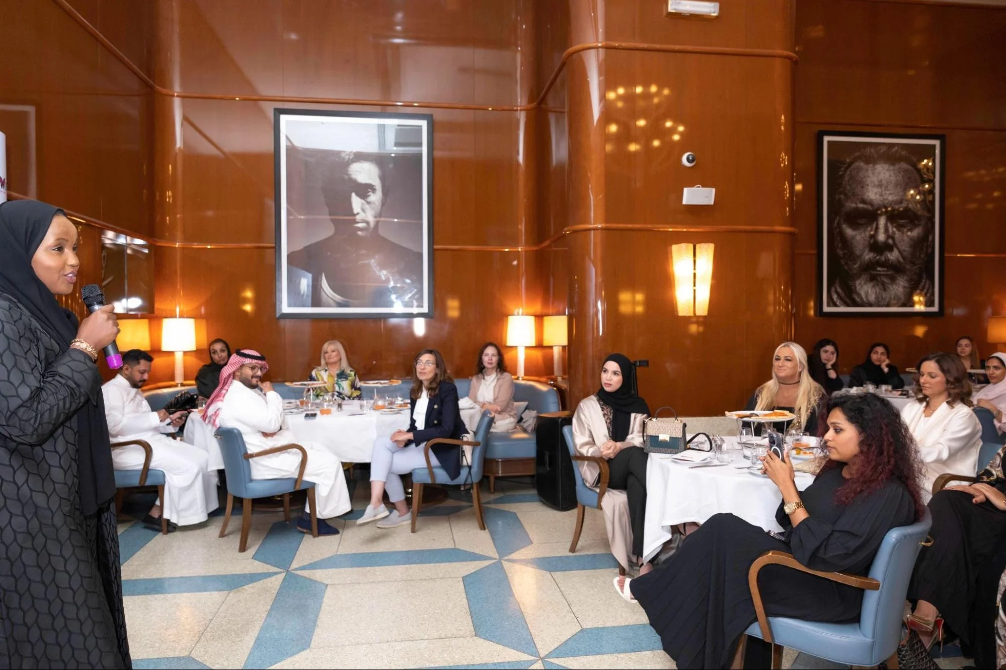 A woman wearing a black headscarf and dark patterned dress speaking into a microphone in a decorated restaurant with large framed black-and-white portraits on the wooden walls and a diverse group of people seated at tables, listening attentively.
