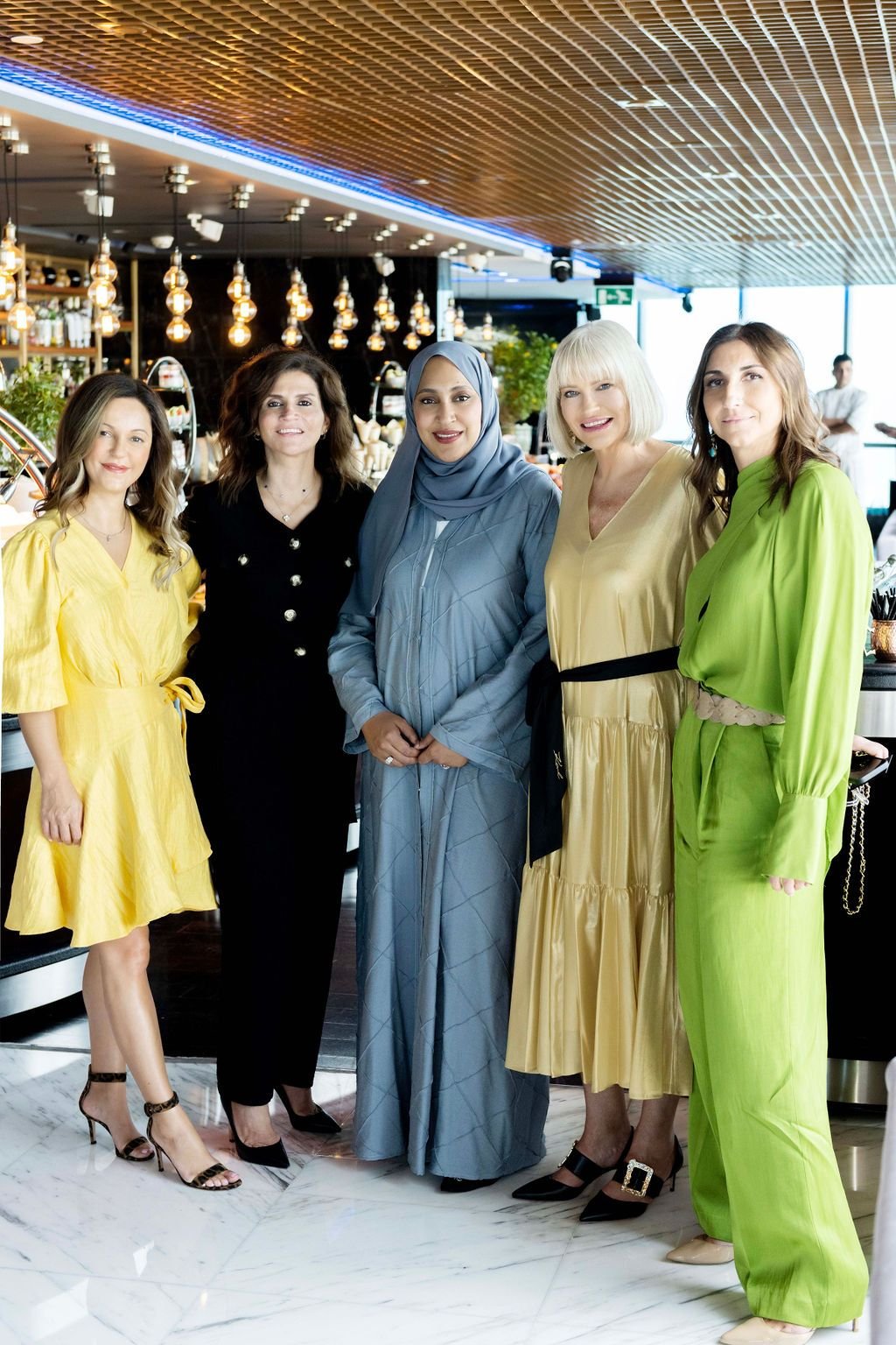 Group of five women standing together in a restaurant, dressed in colorful dresses, smiling for the photo.