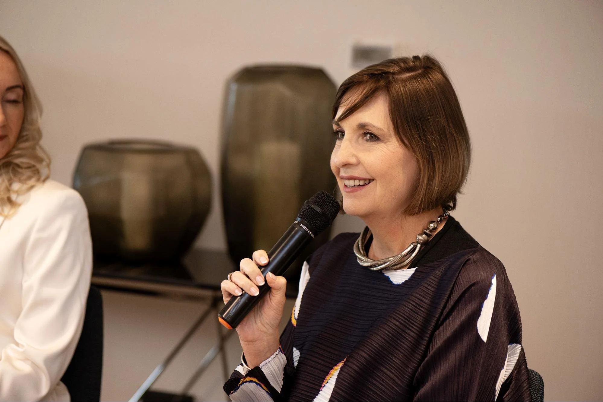 Woman with short brown hair holding a microphone and smiling, sitting in a room with a beige wall and black vases in the background.