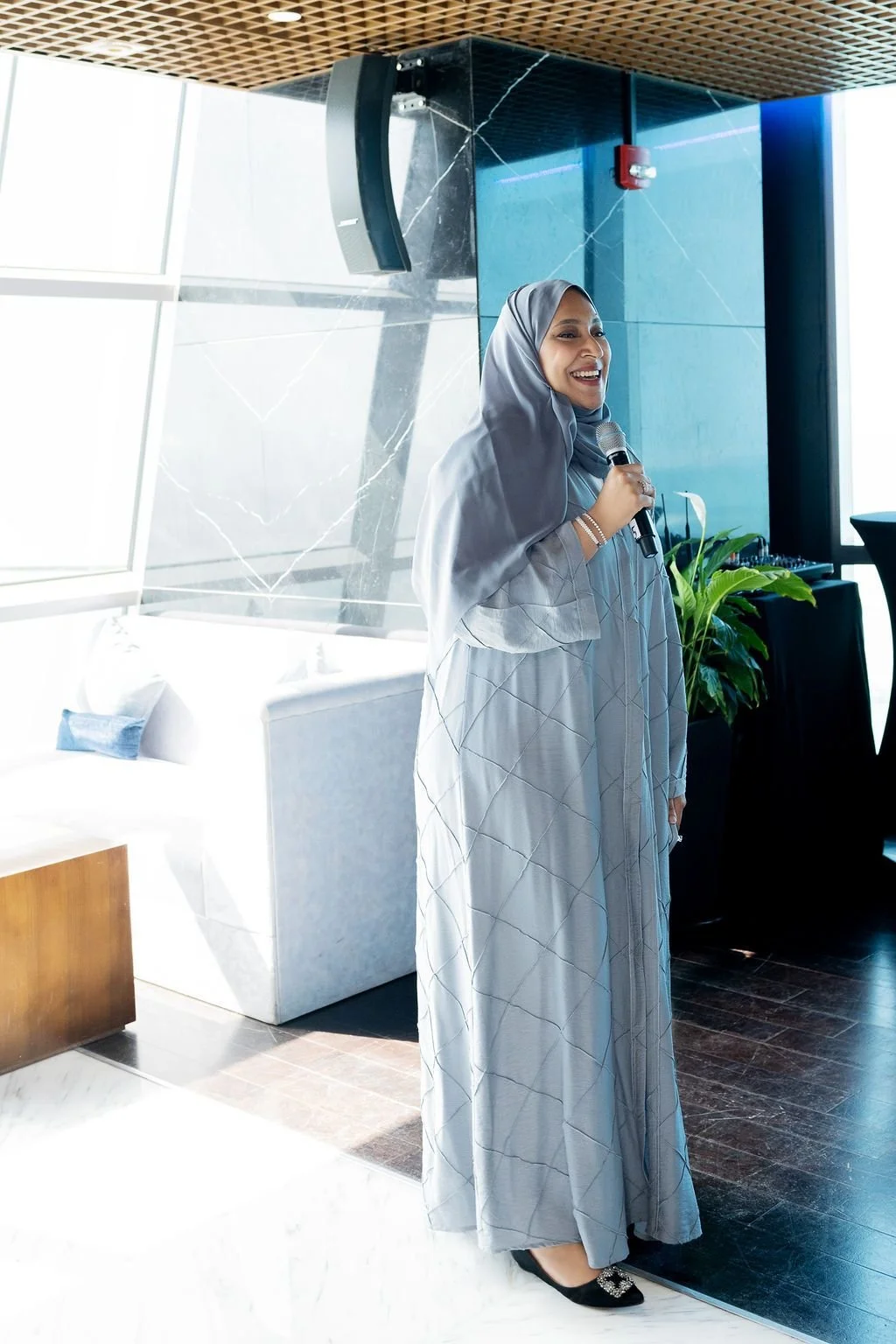 A woman in a gray dress and hijab holding a microphone, standing indoors near a large window with natural light, a white sofa, and green plants in the background.