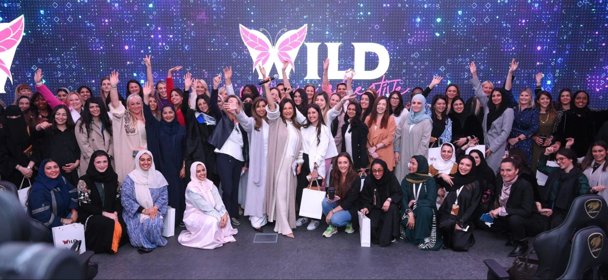 A large group of women posing for a photo at an event, with a vibrant digital background that reads 'WILD' and features a butterfly logo.