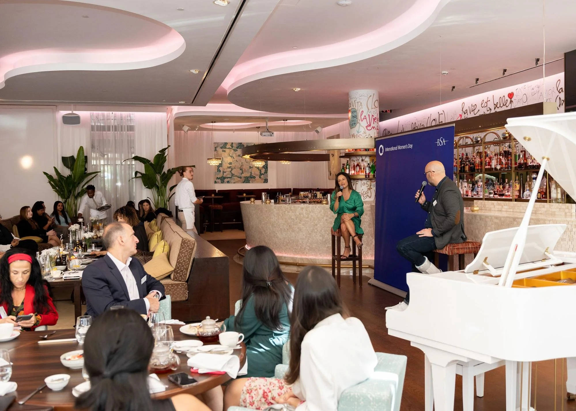 A group of people are seated at tables in a restaurant or event space, attentively listening to a woman speaking into a microphone. A man is seated nearby with a microphone, possibly hosting or participating in a discussion. The room has modern decor