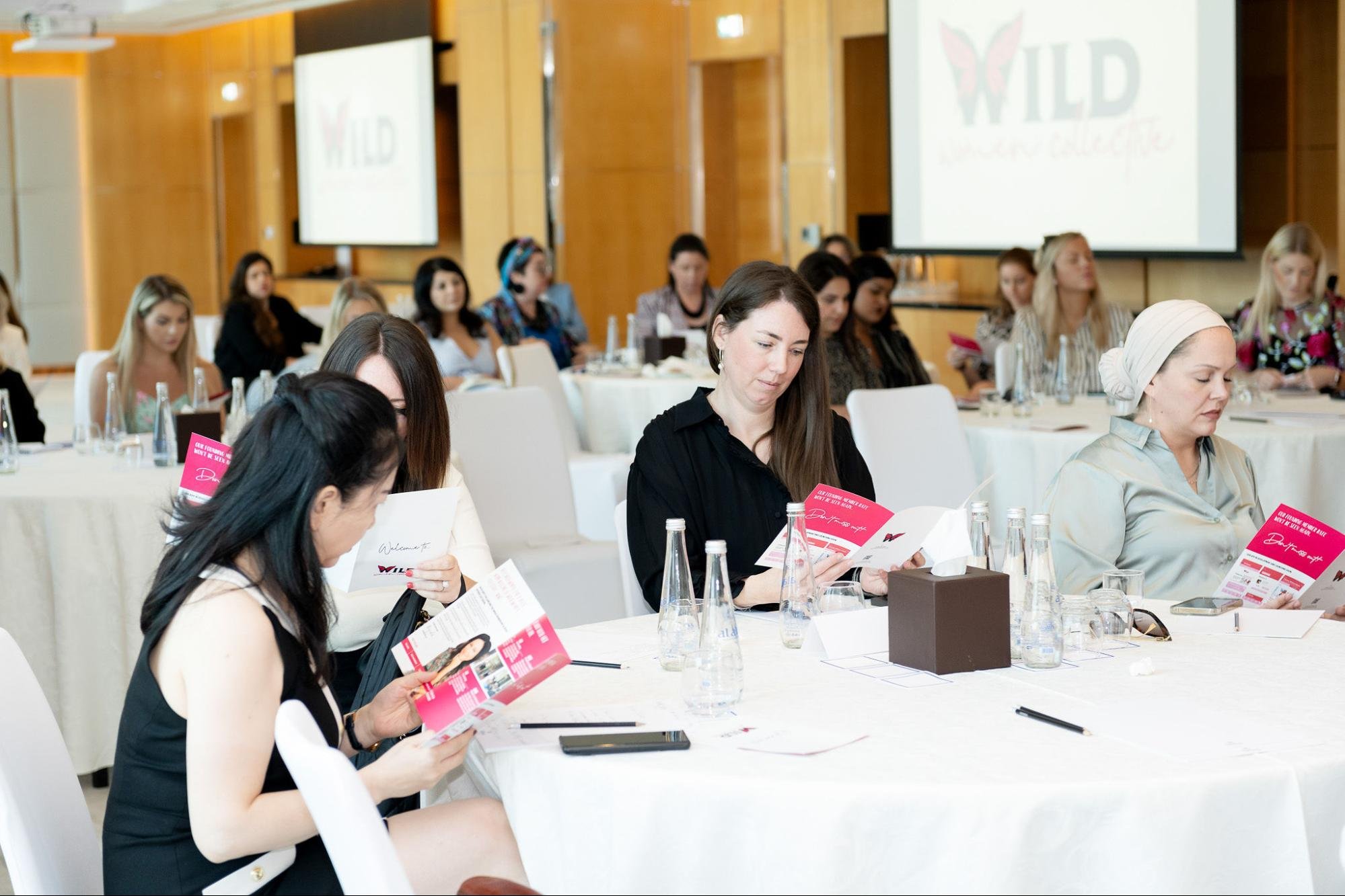 Women attending a conference or seminar, sitting at round tables, reading brochures, with water bottles and notepads, in a room with wooden paneling and large screens displaying logos.