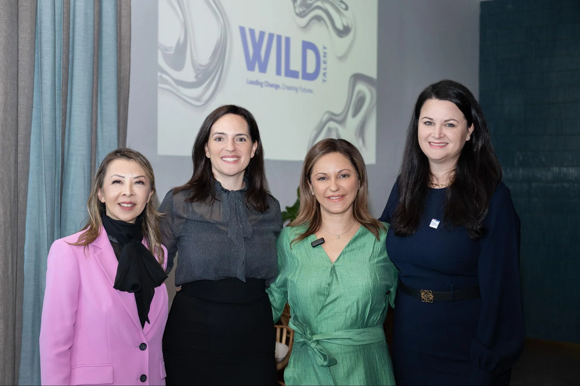Five women standing together at an event, smiling at the camera with a backdrop displaying the 'WILD' logo and slogan 'Leading Change. Creating Impact.'