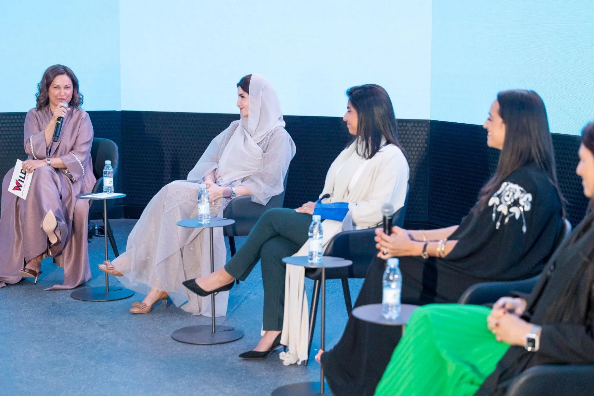 A panel discussion with five women seated in a row, some holding microphones, on a stage with a blue background. The second woman from the left is wearing a hijab, and the others are dressed in varied professional attire.