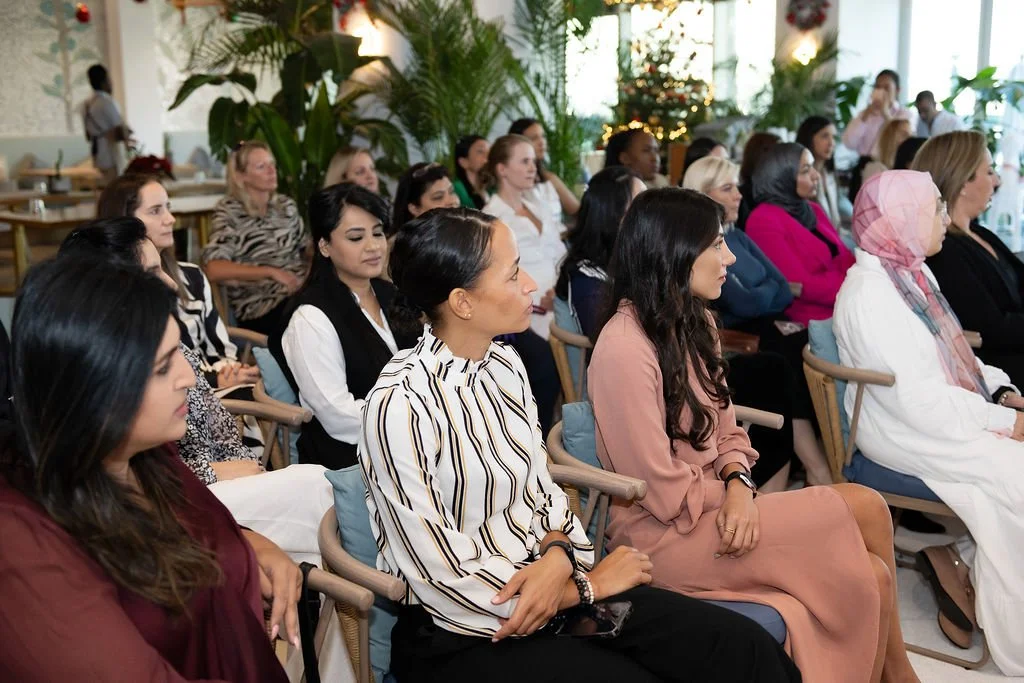 Women seated in an audience at an indoor event, with some wearing headscarves and others in professional attire, surrounded by green plants and festive decorations.