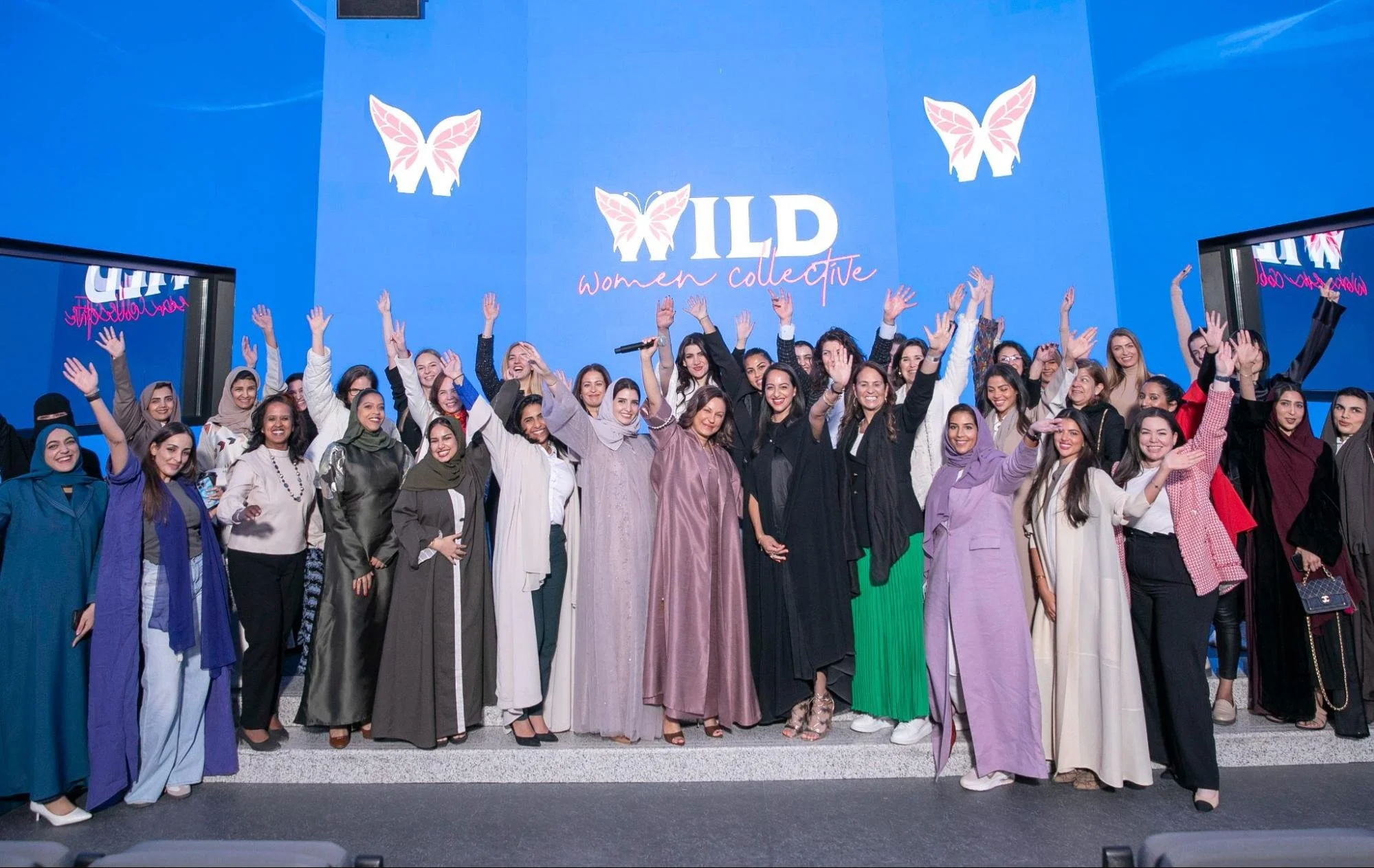 A diverse group of women celebrating at an event called 'WILD women collective,' with many of them raising their hands and smiling. They are standing in front of a blue wall with butterfly and text decorations.