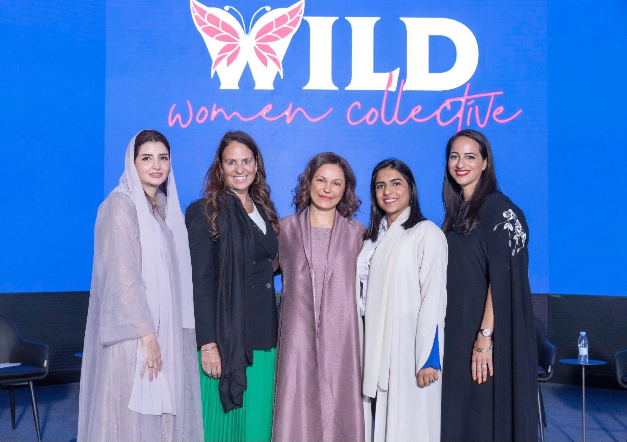 Five women standing together in front of a blue background with the words 'WILD women collective' and a butterfly logo. They are dressed in professional attire and smiling.