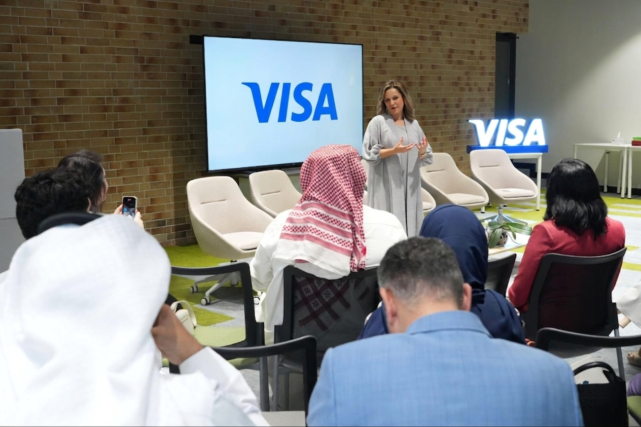 A speaker presenting at a Visa event, with an audience of diverse individuals, some wearing traditional Middle Eastern attire, seated and listening attentively in a modern conference room with Visa branding displayed on screens.