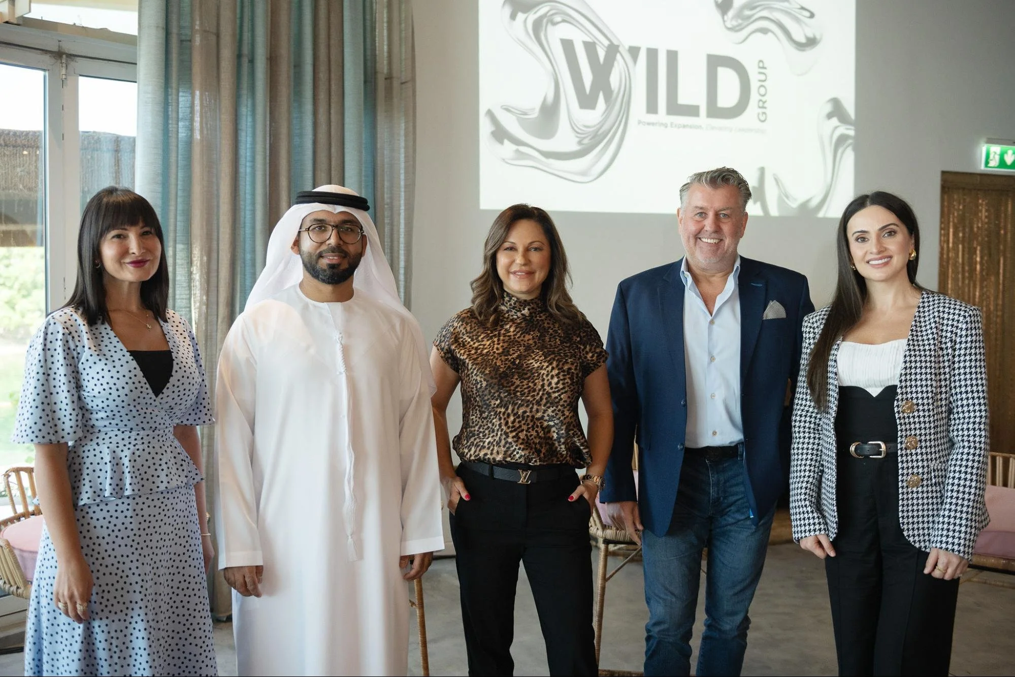 Five diverse adults standing together indoors at a professional event. They are smiling and dressed in business casual attire. The background features a window, curtains, and a large screen displaying the logo 'WILD GROUP' and the tagline 'Powering Expansion, Creating Trust'.