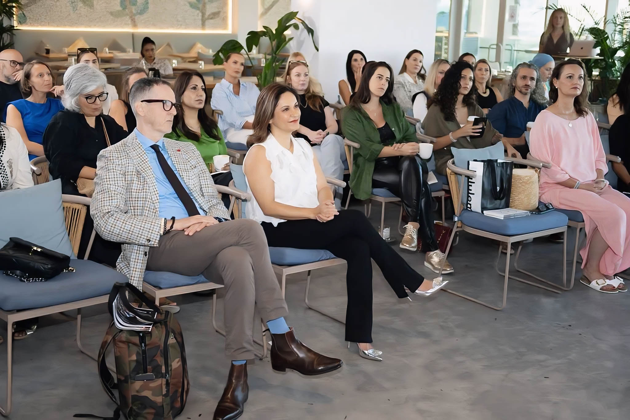 A group of diverse people sitting and listening during a conference or seminar in a modern bright room with large windows and greenery.