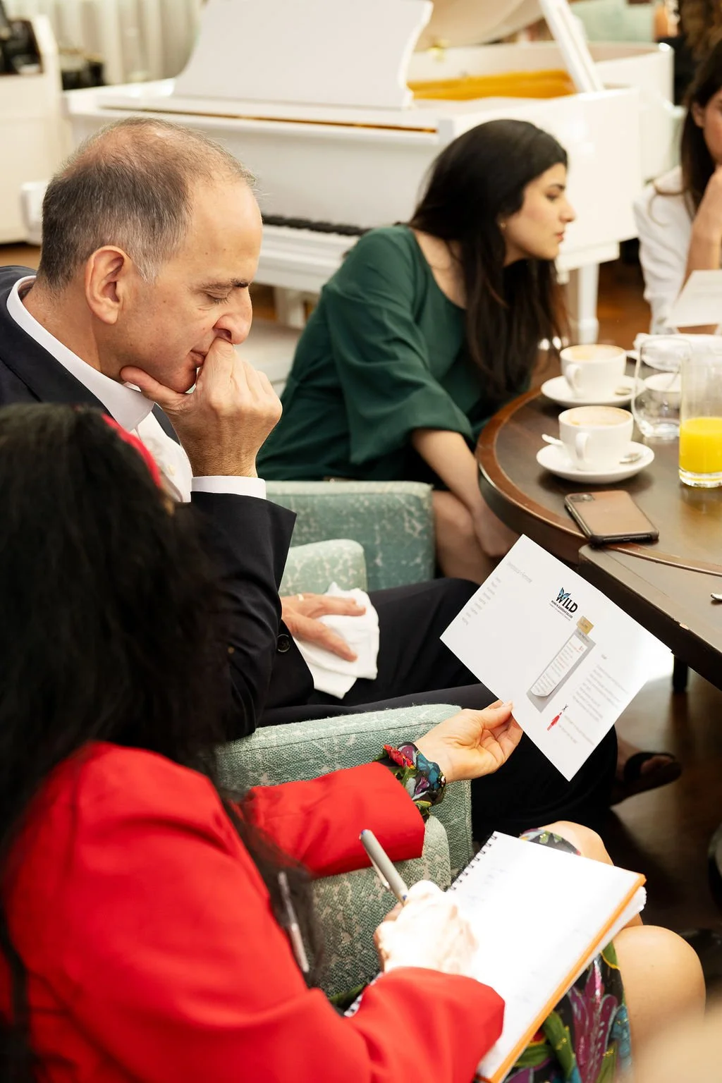 A group of people sitting at a table during a meeting or discussion, with cups of coffee, a glass of orange juice, and various papers and notebooks on the table.
