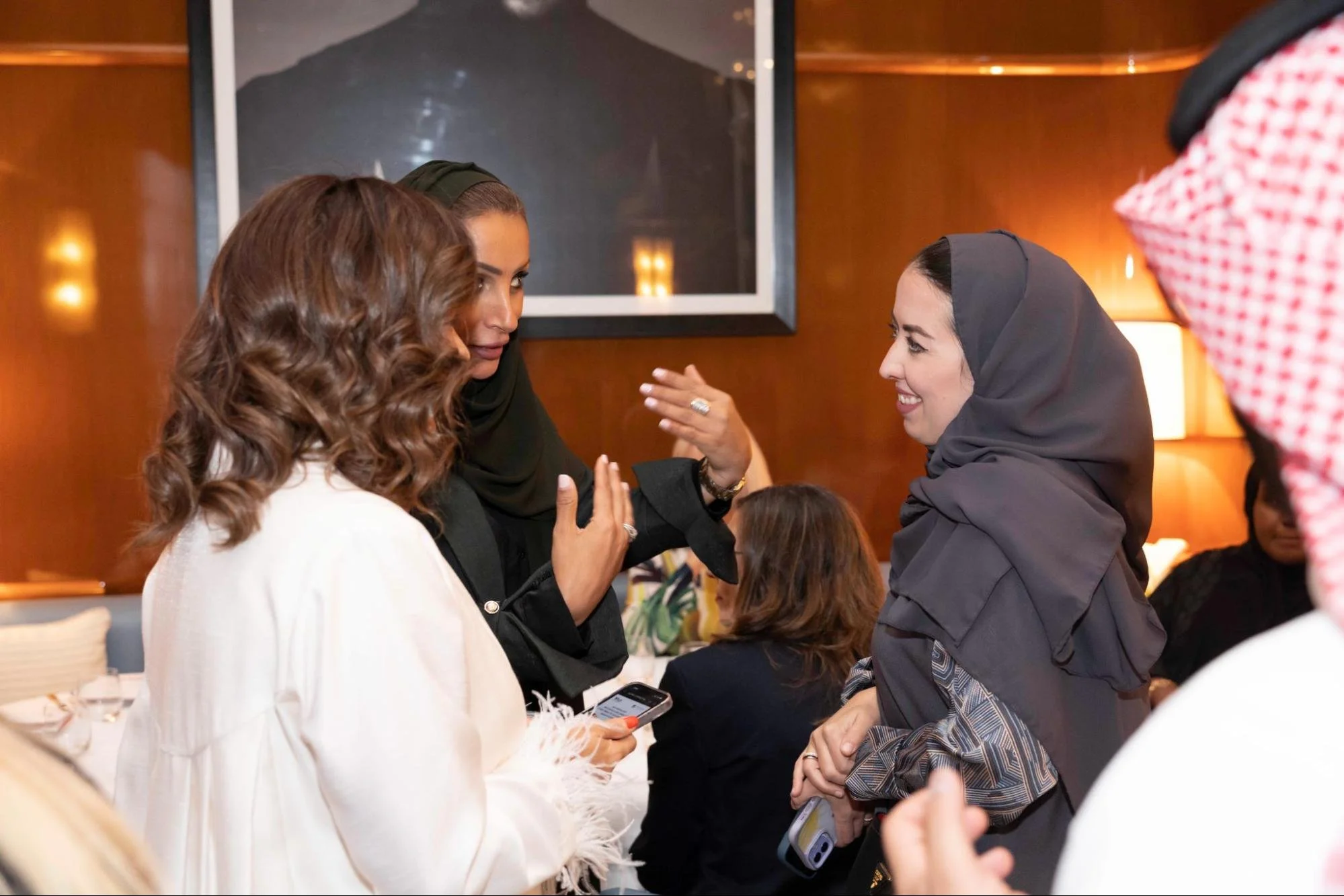 A group of women engaging in conversation at a social gathering. Two women are smiling and gesturing with their hands. The woman on the left has wavy brown hair and a white outfit, while the woman on the right is wearing a dark headscarf and a patterned garment. In the background, other people are seated at a table in a warmly lit room with wooden walls.
