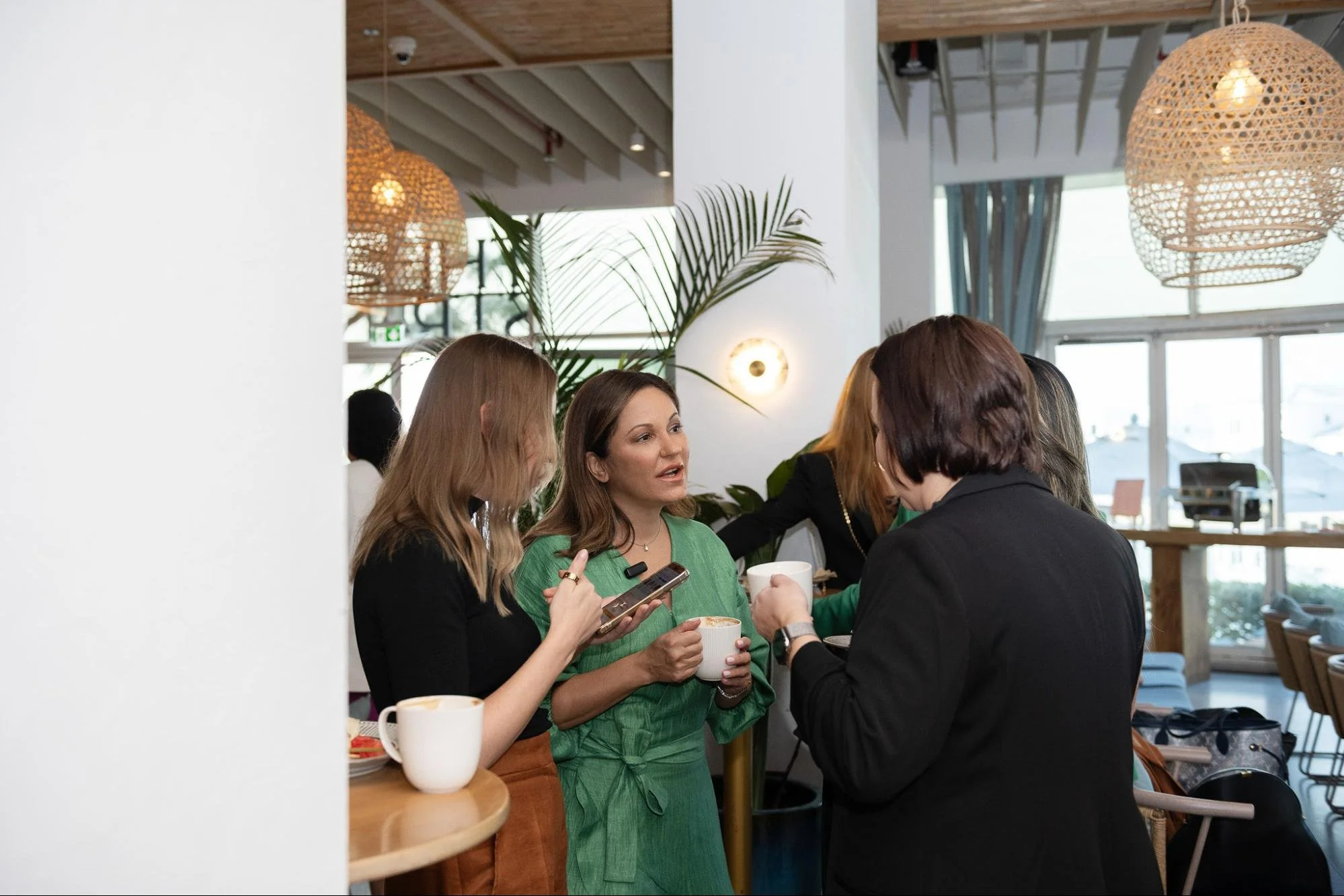A group of women in a cafe engaged in conversation. They are holding cups of coffee and talking to each other. The cafe has large windows, hanging wicker lamps, and various plants.