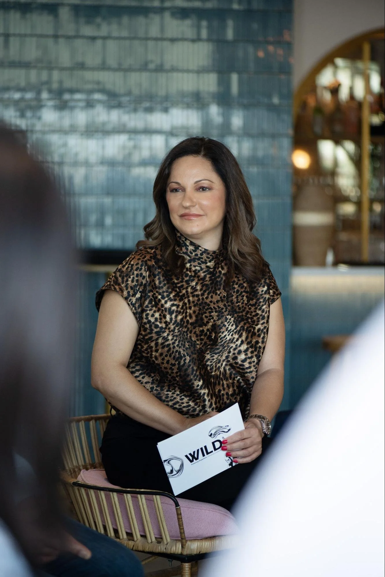 A woman with long but wavy brown hair wearing a leopard print top and holding a brochure that says "WILD" sits on a pink cushioned chair in a restaurant or cafe. She is smiling slightly and appears to be engaged in conversation with someone off-camera.