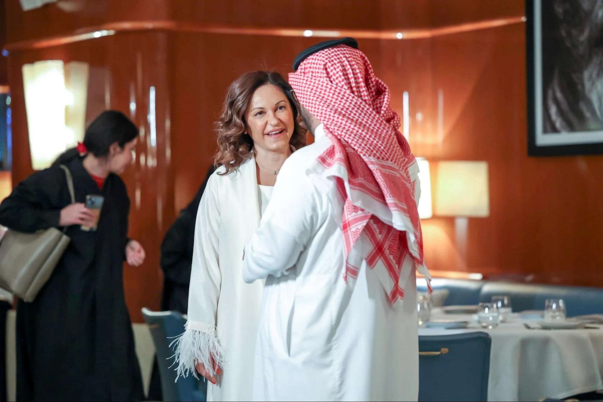 A woman in a white blouse interacts with a man wearing traditional Middle Eastern attire, including a red and white keffiyeh, in a warmly lit setting with other people in the background.