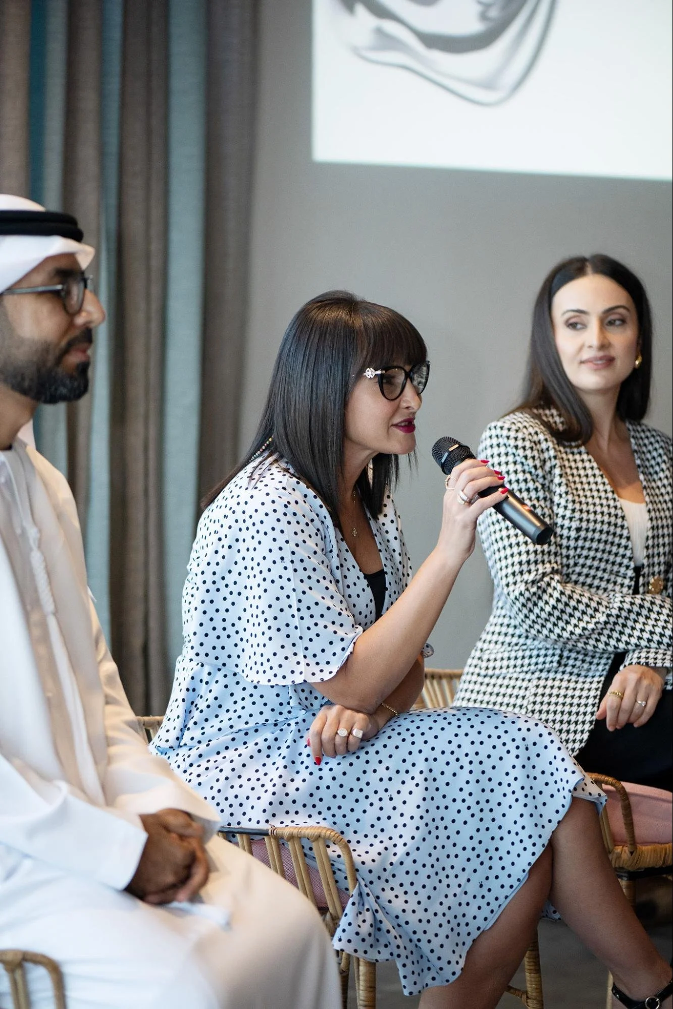 Three individuals seated at a panel discussion or meeting. The woman in the middle is speaking into a microphone, wearing glasses and a white dress with black polka dots. The woman on the right is looking at her with a slight smile, dressed in a matching black and white outfit. The man on the left is wearing traditional Middle Eastern attire, including a white thobe and head covering. The setting appears to be a conference or seminar room.