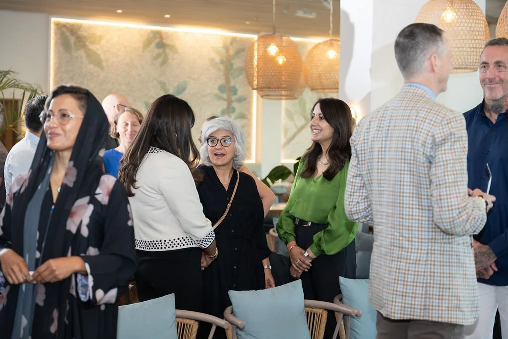 Group of people conversing and smiling at a social gathering indoors. The setting has large woven pendant lights and leafy wall decor.