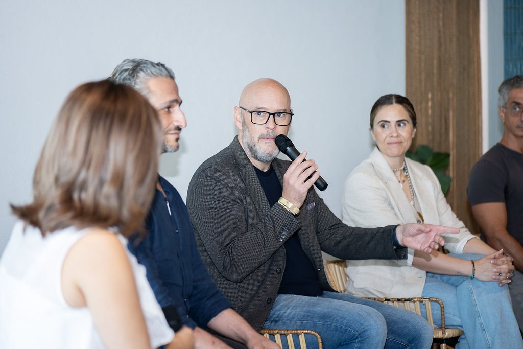 Group of five people seated during a panel discussion, one man speaking into a microphone, in a professional setting.