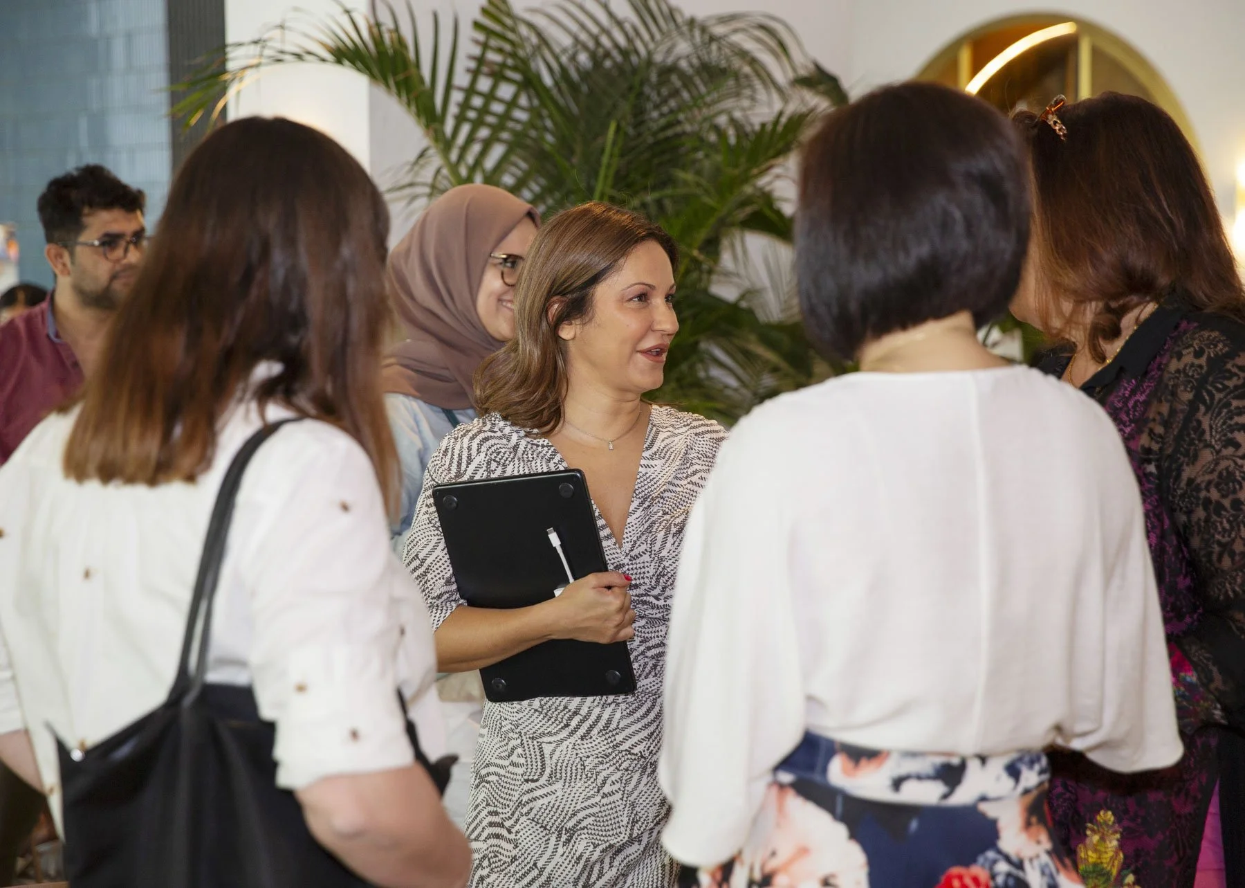 Group of women engaged in conversation, with one woman holding a clipboard, in an indoor setting with greenery in the background.