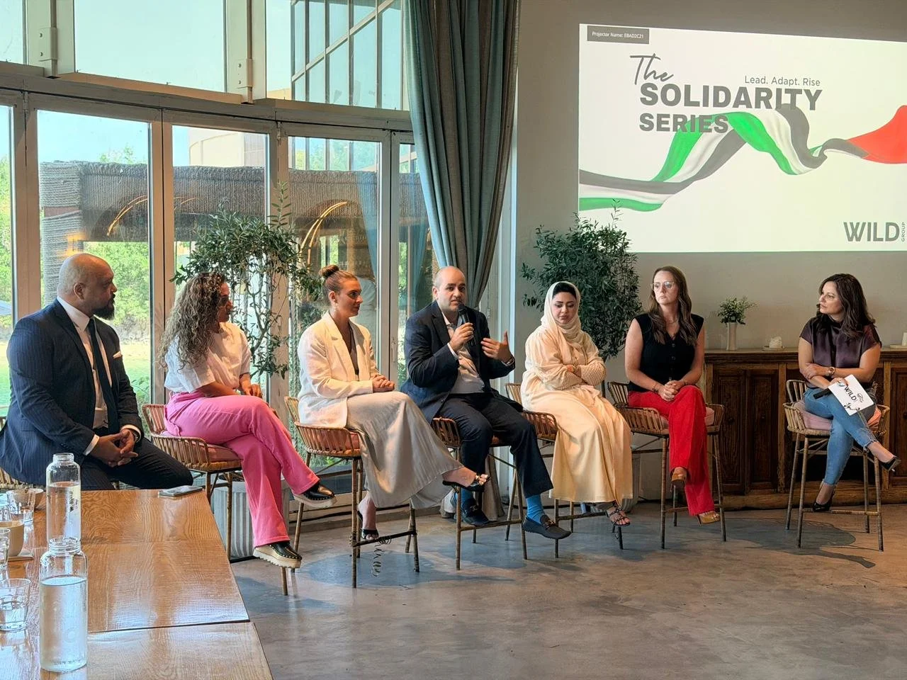 A panel of seven people sitting on chairs during a discussion event. The backdrop shows a large screen with the text 'The Solidarity Series' and a graphic with green, white, black, and red colors. The setting has large windows, potted plants, and a wooden table with water bottles in the foreground.