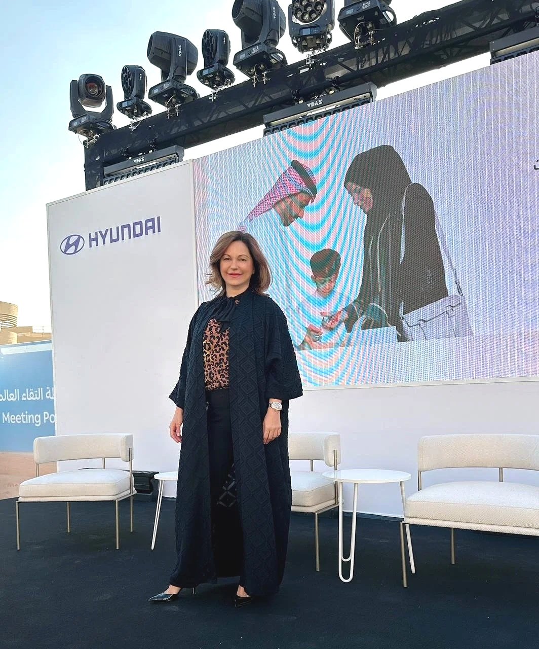 A woman standing on a stage at an event, with a large screen behind her displaying the Hyundai logo and an image of two people exchanging business cards.