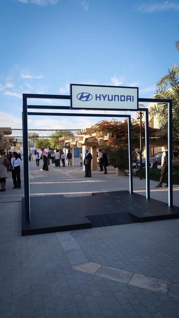 Hyundai sign at the entrance of an outdoor event or exhibition area with people gathered, some standing and talking, against a clear blue sky.