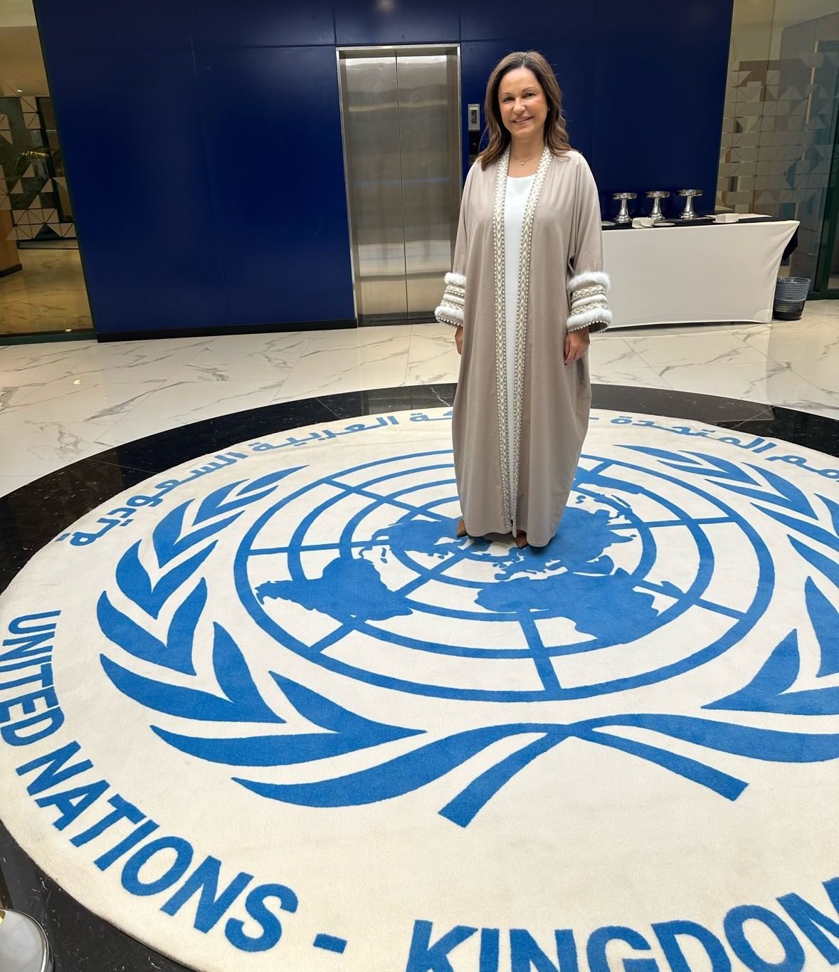 Woman standing on a UN logo rug in a lobby with blue and beige walls, wearing a beige abaya with white and gold details.