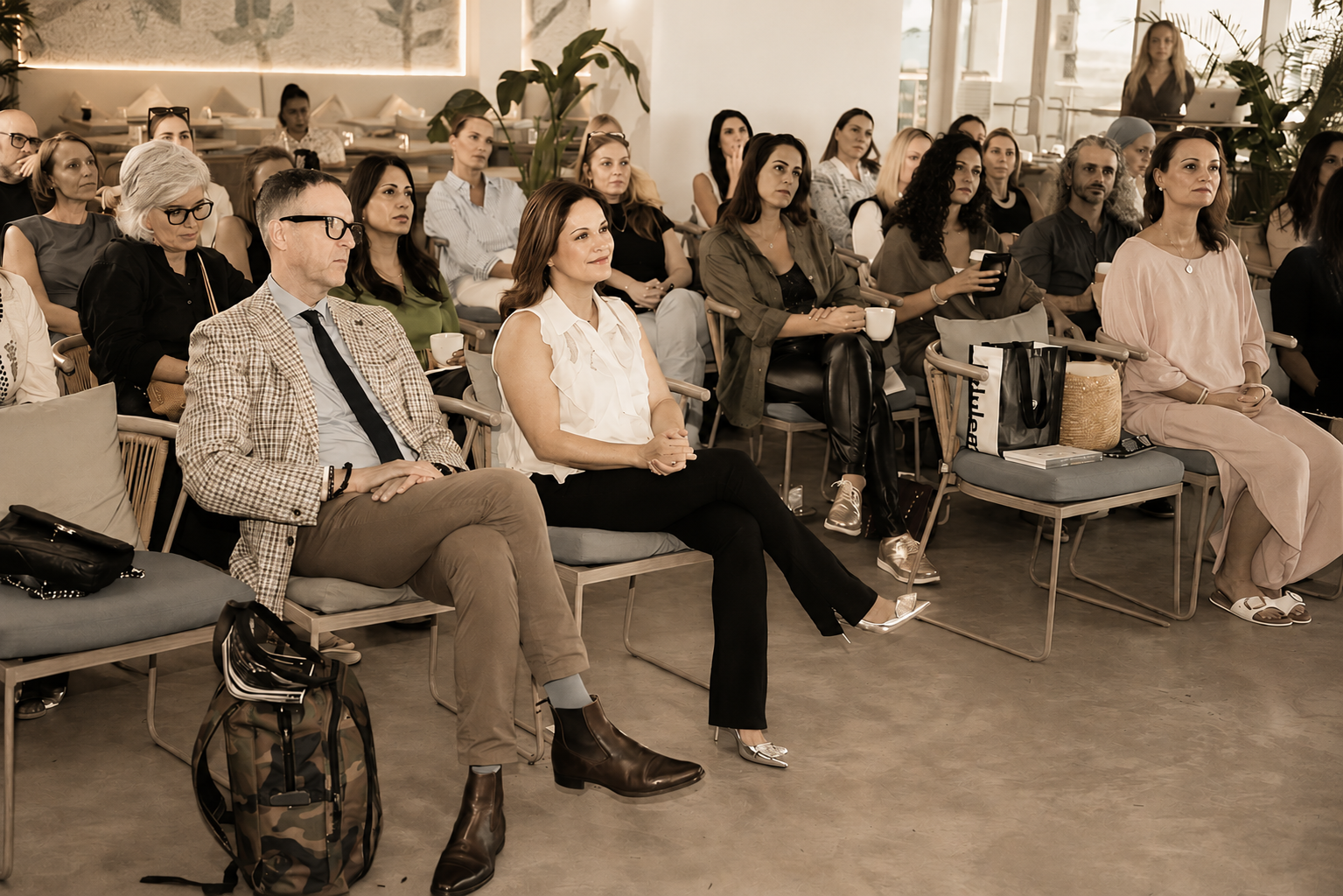 A diverse group of people seated and attentively listening at an indoor event or seminar, with some holding cups, in a modern, well-lit room with plants and large windows.