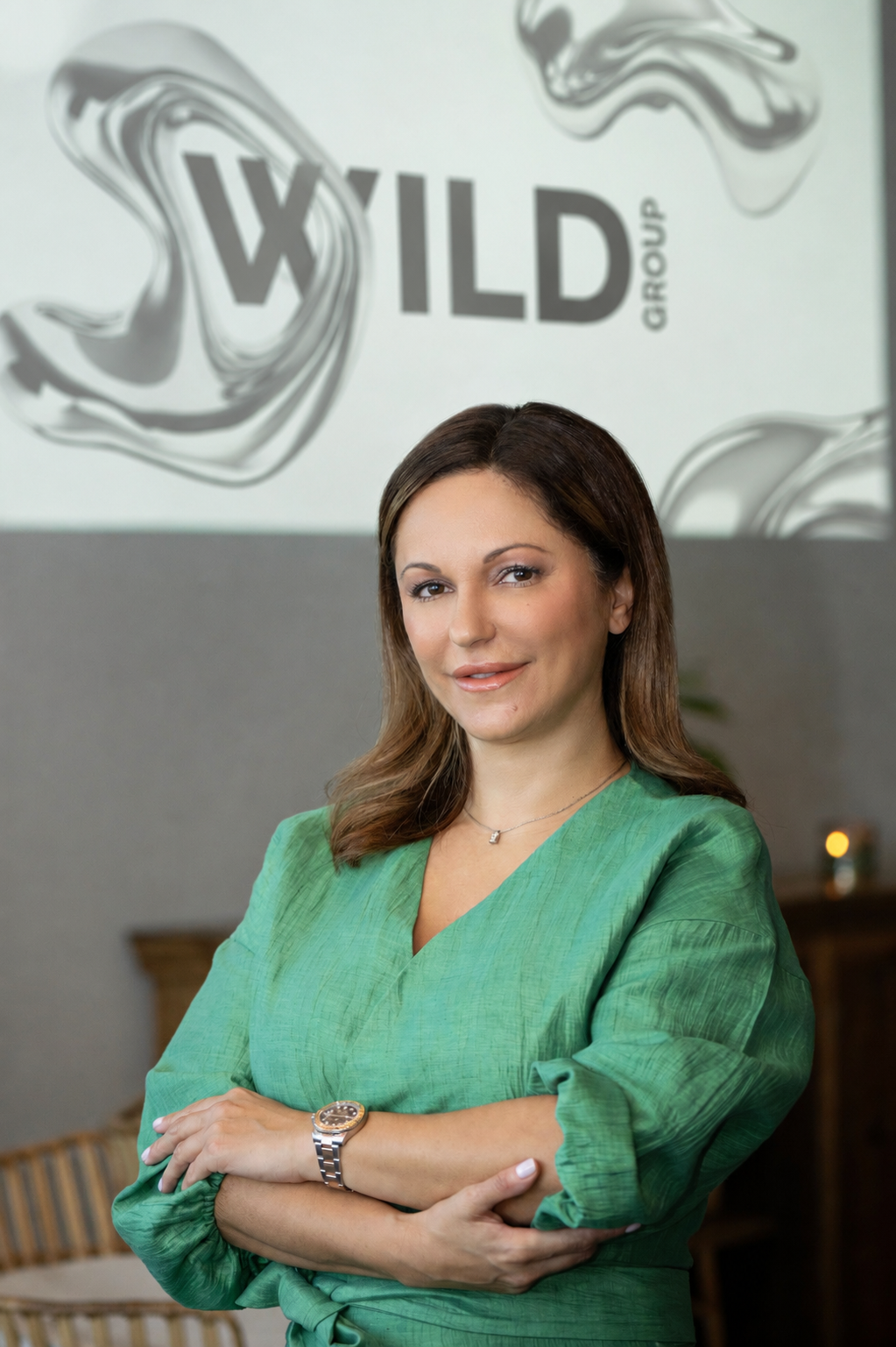 A woman in a green dress with crossed arms, standing indoors against a background featuring the 'Wild Group' logo projected on the wall.