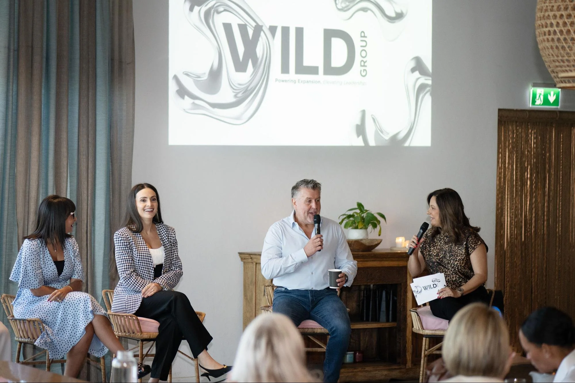 Group of people seated and standing indoors at a panel or discussion event. There is a man with a microphone speaking, and three women are smiling and listening. A projected sign with the logo 'WILD GROUP' is on the wall behind them. Decorative curtains, a wooden mantel, and a plant are in the background.