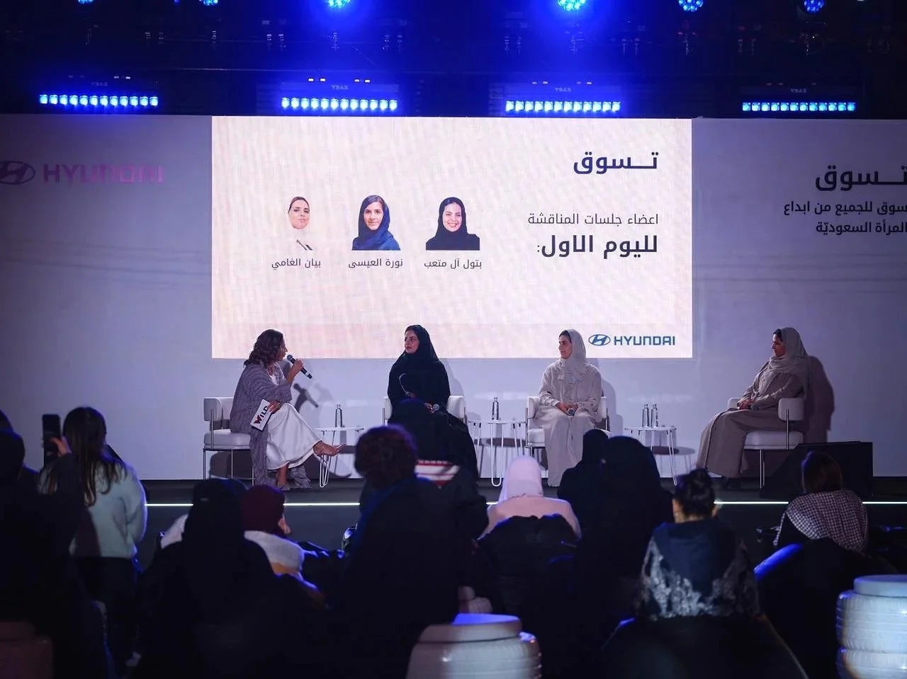 Women sitting on stage during panel discussion at an event, with large screen behind displaying event details in Arabic and photos of three women with headscarves, branding for Hyundai.