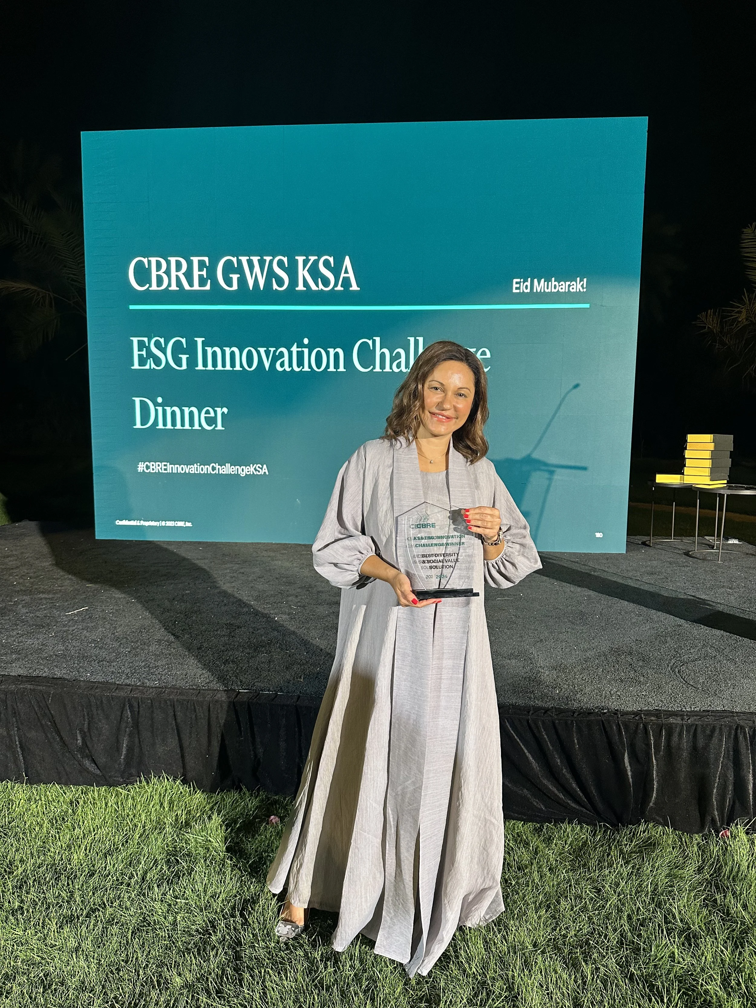 A woman standing on grass at night holding a clear award plaque, with a large blue event banner behind her that reads 'CBRE GWS KSA', 'Eid Mubarak!', and 'ESG Innovation Challenge Dinner'.