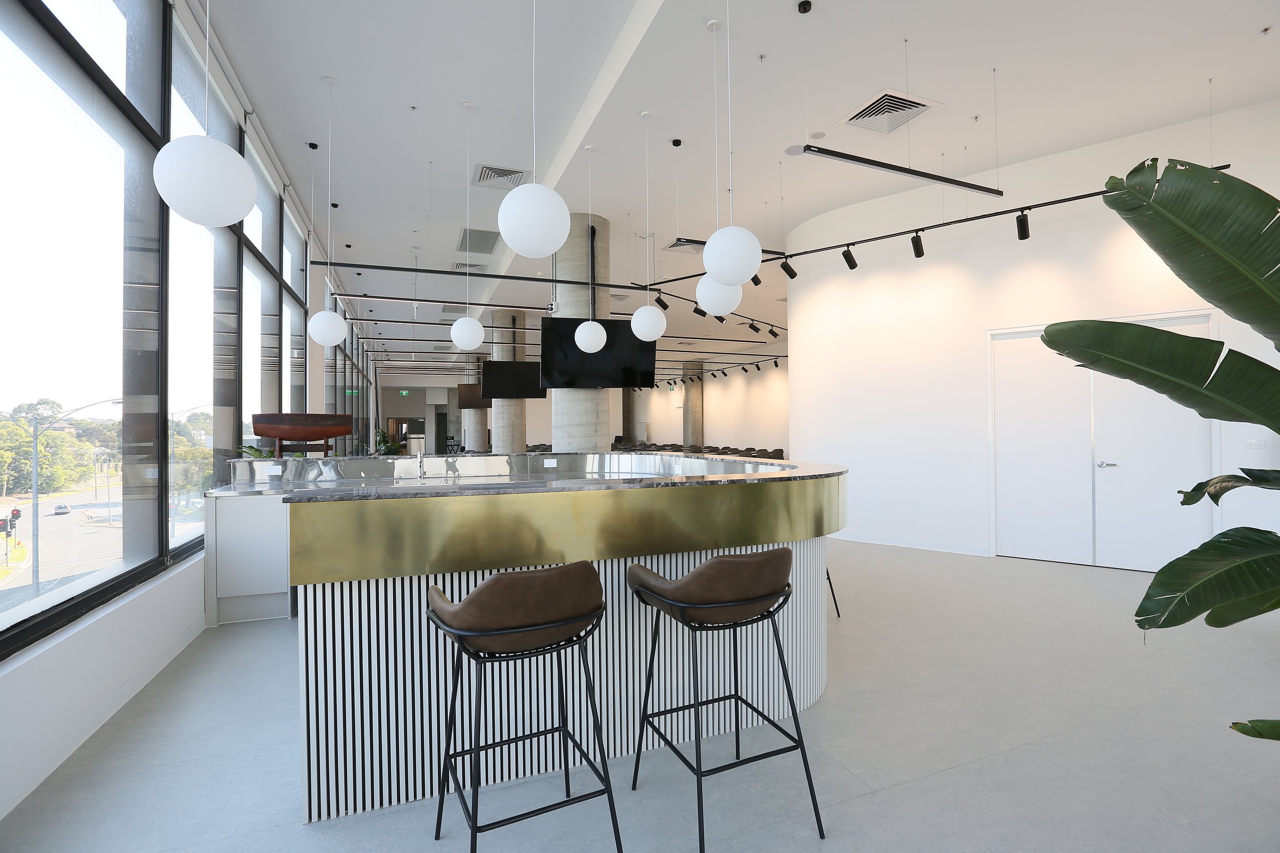 Modern interior space with large windows, hanging white globe lights, a curved bar with a gold front, high chairs, and large green leaves in the foreground.