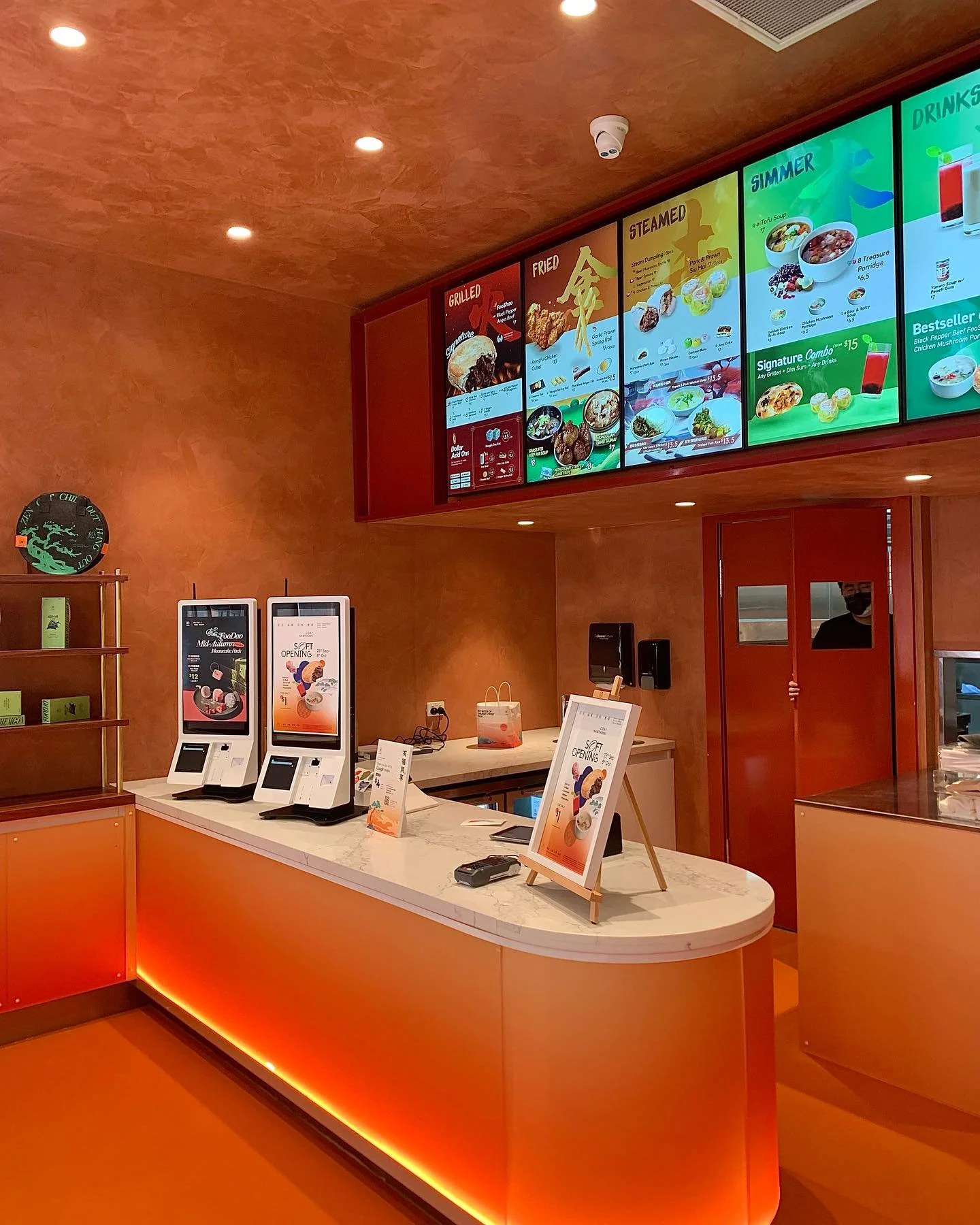 Counter at a restaurant with two self-service ordering kiosks, a signboard on a small easel, and digital menu screens above the counter showing menu items and prices. The interior has orange walls and a marble countertop.