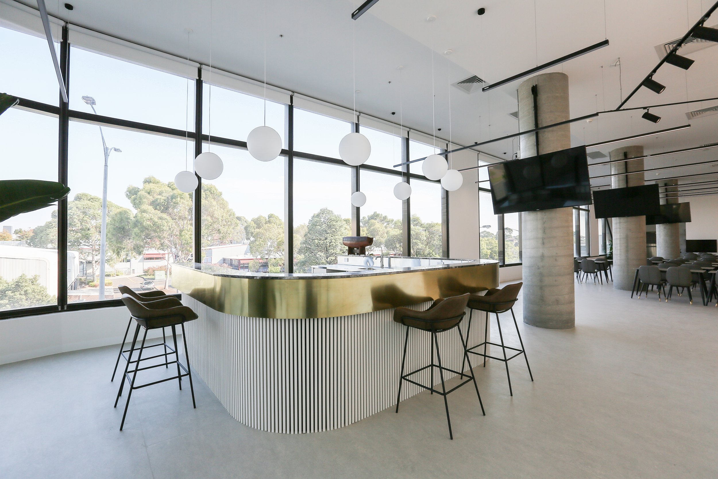 Modern interior space with large windows, hanging spherical light fixtures, a gold-colored curved bar or counter, chairs, and a flat-screen TV, overlooking trees outside.