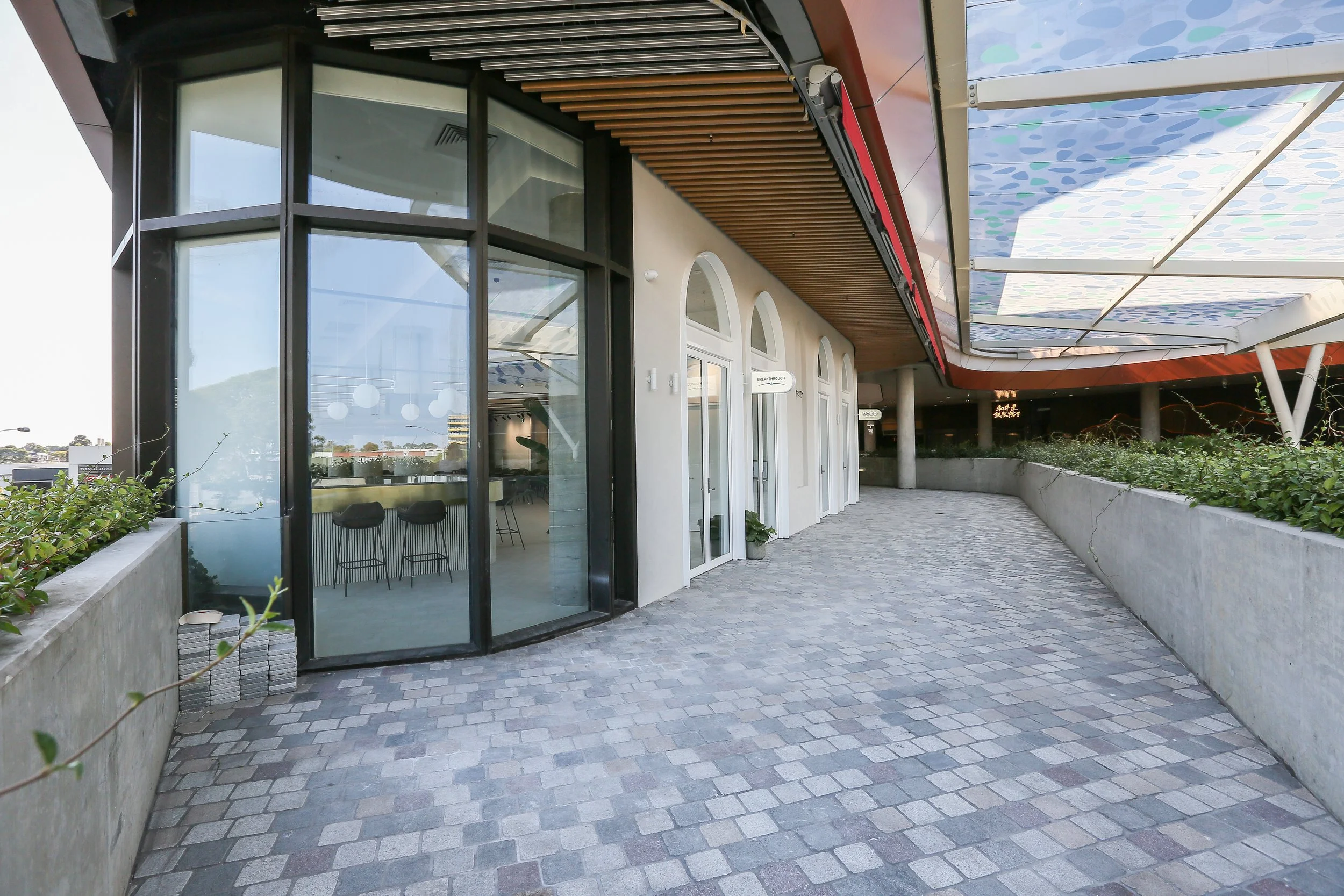 Empty outdoor walkway with gray stone pavers, concrete planters with green plants, glass doors to a building, and a decorative overhang with trees and sky reflected on the glass.