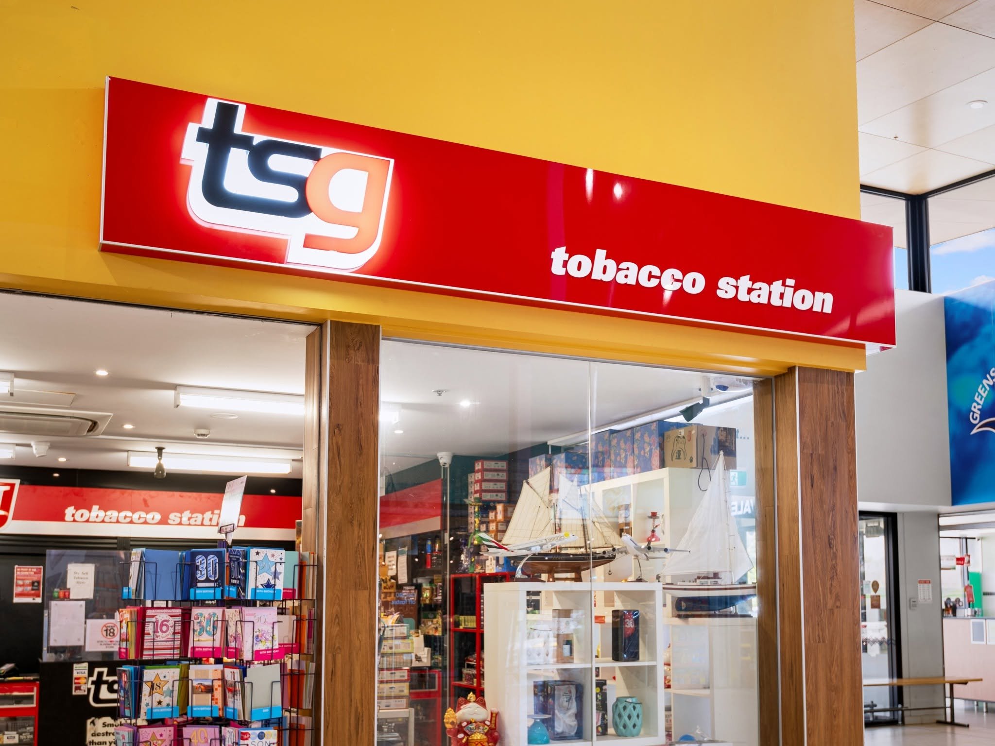 Sign for tobacco station inside a shopping mall or store, with shelves displaying greeting cards and model airplanes in the background.