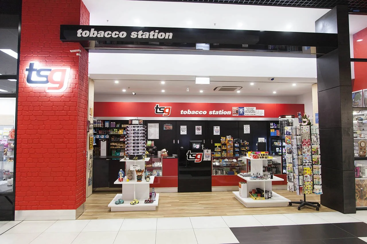 Inside a tobacco shop with a red and black color theme. The shop has shelves with tobacco products, accessories, and gift items. Signage at the top reads 'tobacco station' and the store logo shows 'tsg'. There are display tables with small items and 