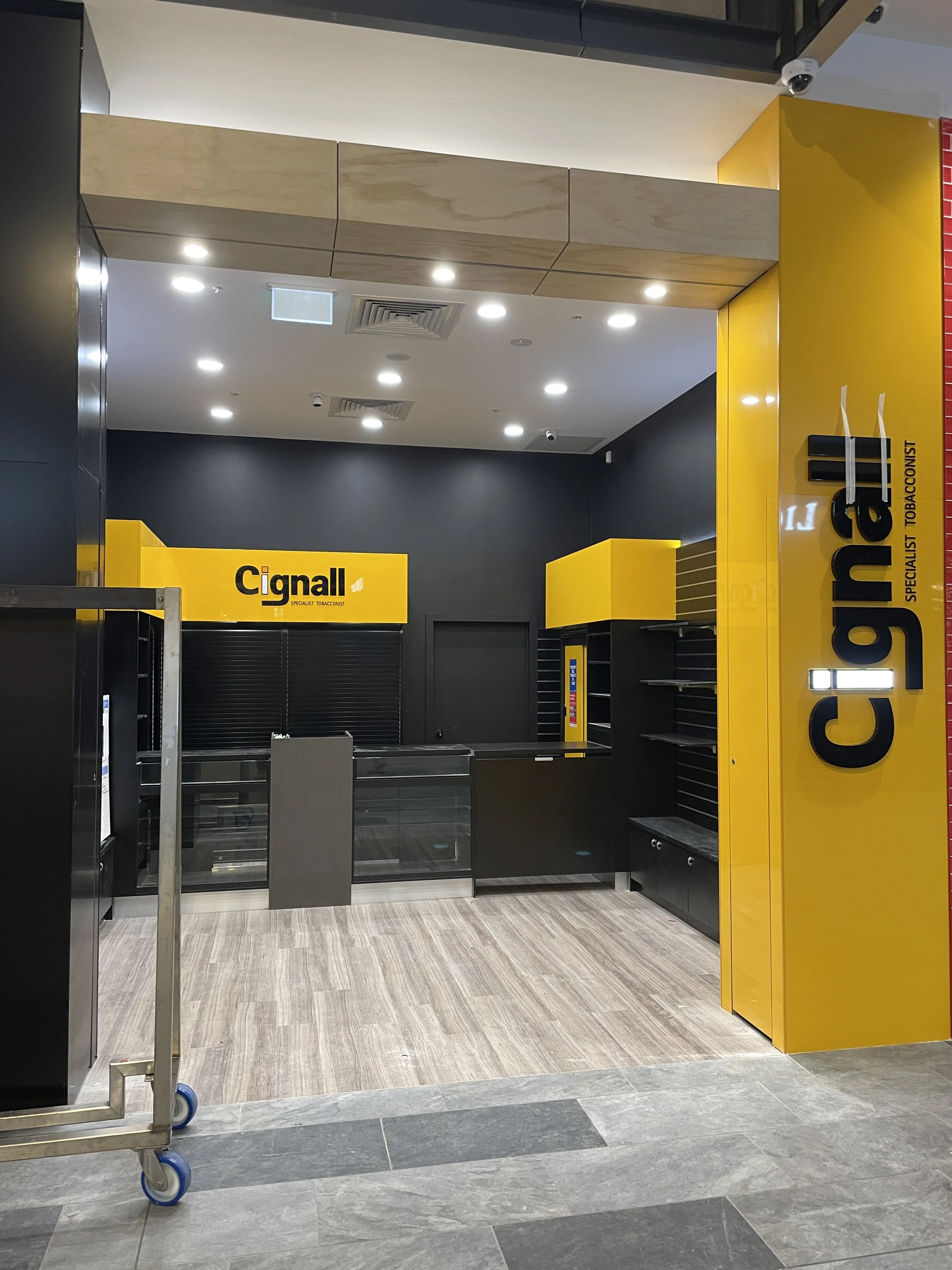 Empty retail store with yellow signage that reads 'Cignall', black shelves, and a shopping cart with blue wheels in front, inside a mall or commercial space.