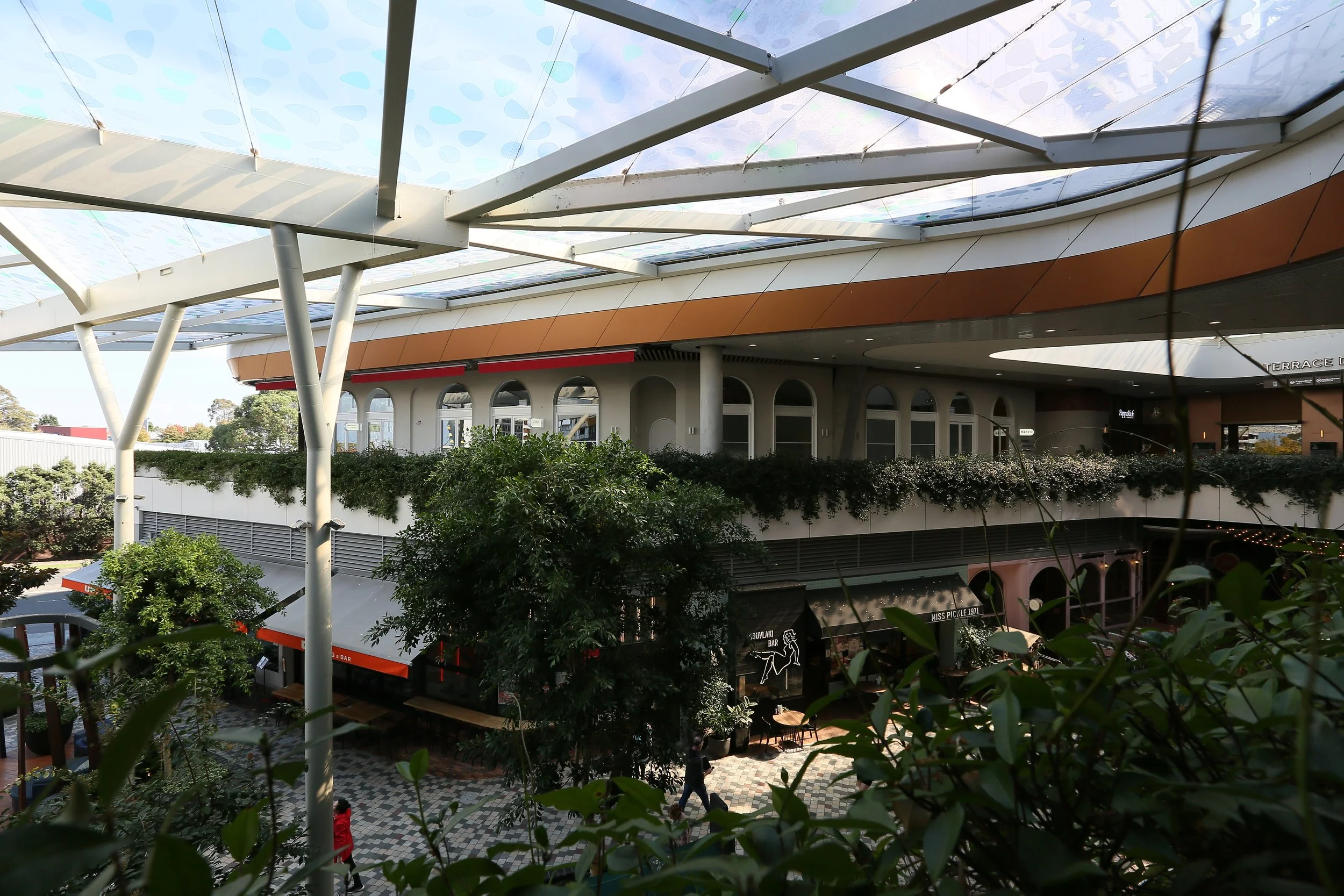 Outdoor shopping or entertainment mall with a curved rooftop covered with glass panels, trees and greenery on a terrace level, and people walking on a checkered pavement below.