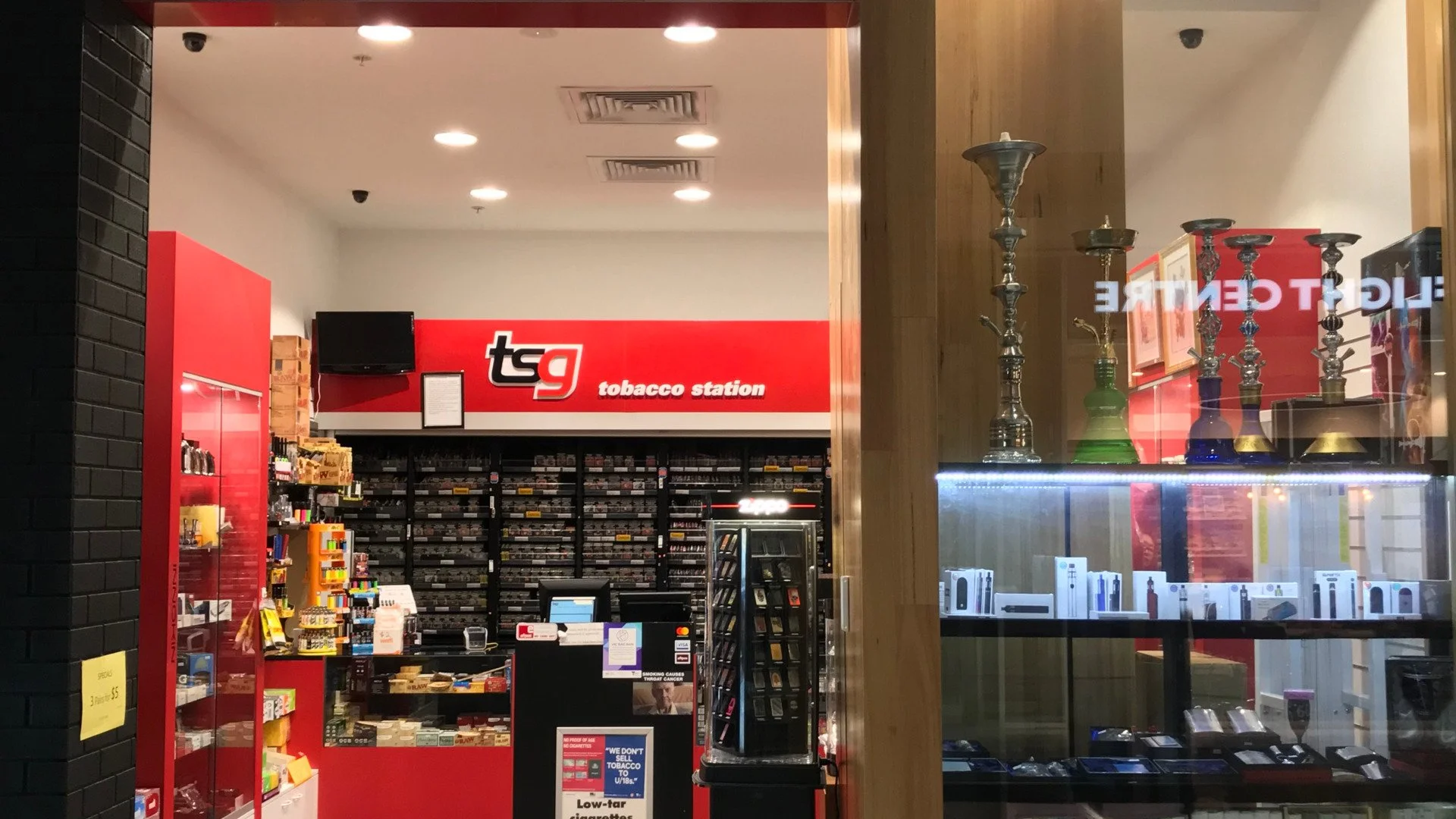 View of a tobacco shop named 'tq tobacco station' with shelves stocked with cigarettes and tobacco products, a cash register, and hookahs displayed on a wooden shelf at the front window.