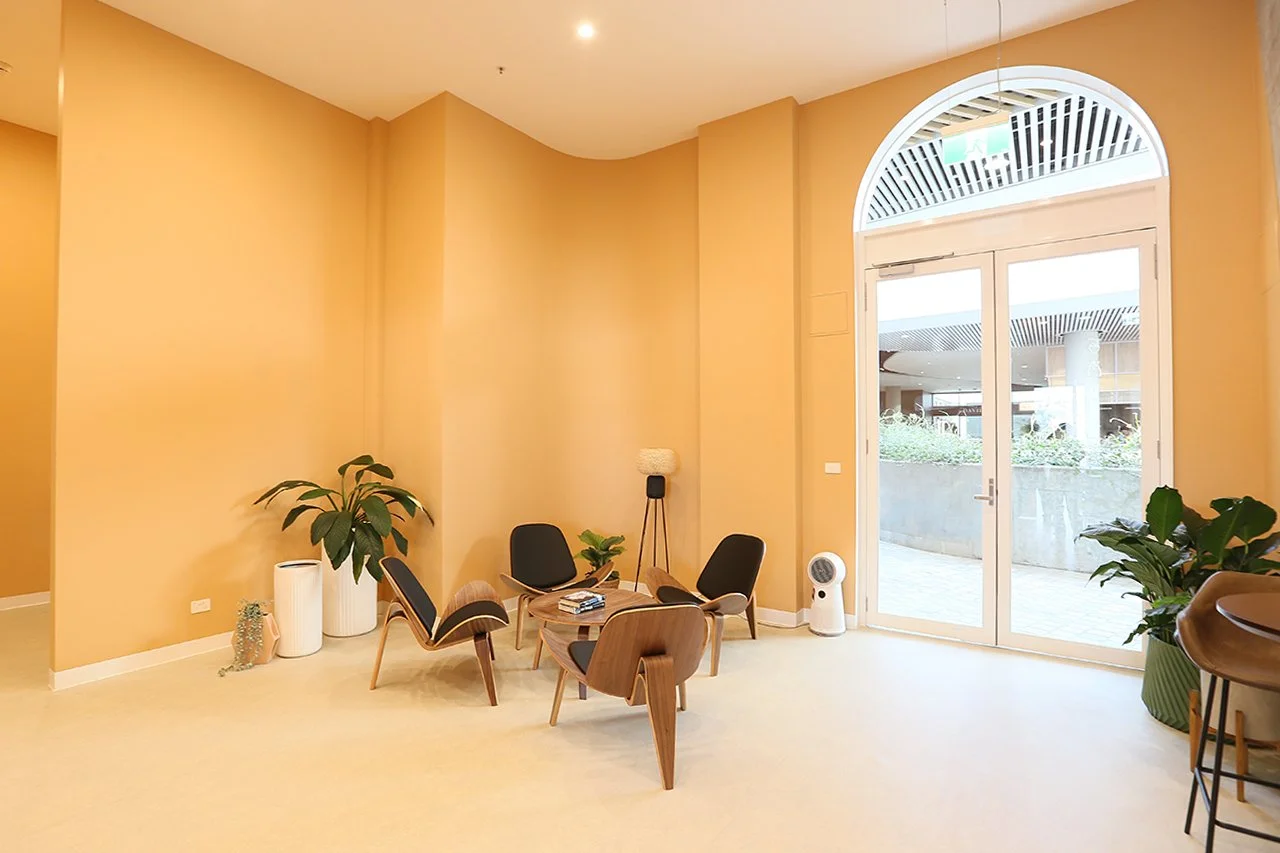 A modern waiting area with four black and wooden chairs around a small wooden table, potted plants, a floor lamp, and large glass doors leading outside.