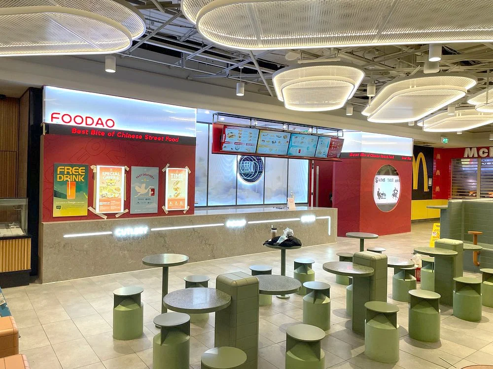 Empty food court area with a Foodao street food restaurant counter, digital menu screens above, and McDonald's visible in the background. There are green round tables and stools arranged throughout the space, with decorative lighting fixtures on the 