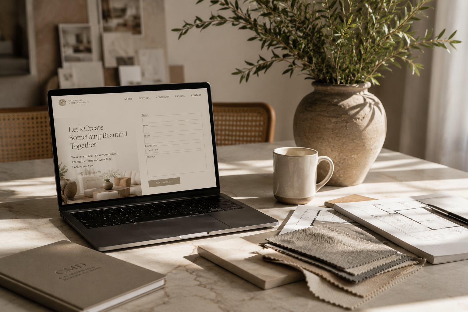 Laptop on a marble table displaying a website with a contact form, surrounded by a ceramic mug, a large vase with green foliage, fabric swatches, a sketchbook, and architectural blueprints in a well-lit room.