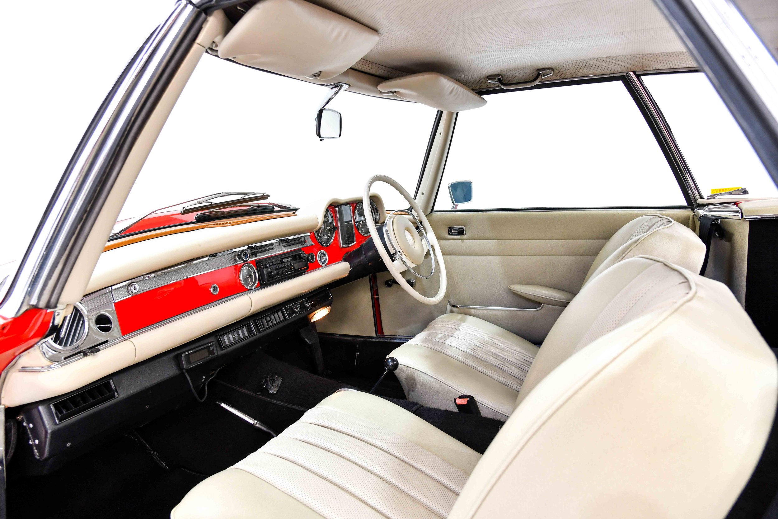 Interior of a vintage car with cream-colored seats, dashboard, and ceiling, featuring a red dashboard with analog gauges, a steering wheel, and side mirrors.