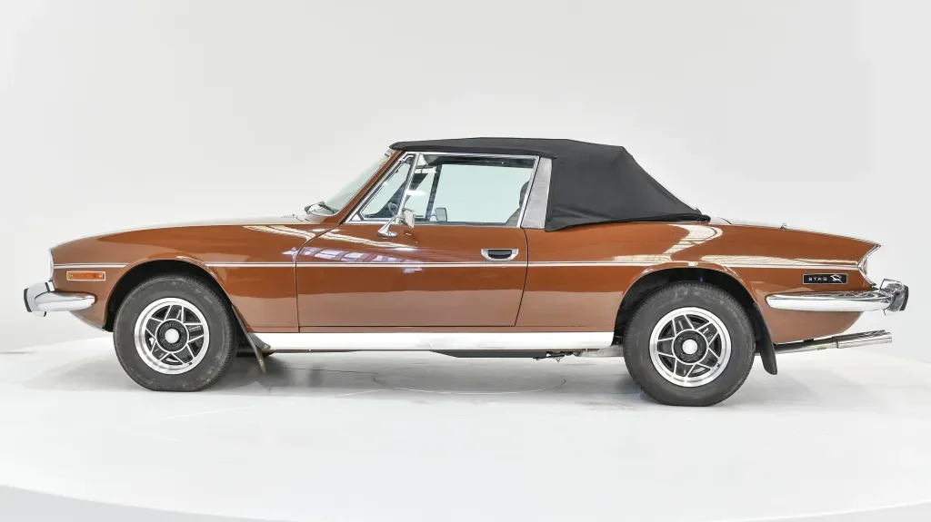 A vintage brown convertible sports car with a black soft top, parked against a plain white background.